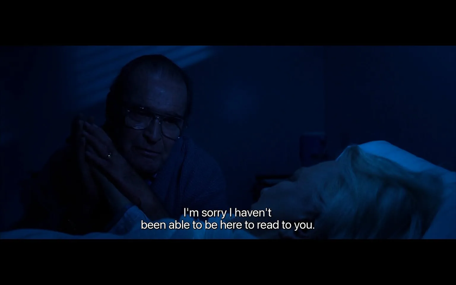 The image depicts a dimly lit indoor setting, likely a bedroom, characterized by a blue hue that suggests nighttime or low lighting. 

In the foreground, there are two individuals. On the left, an older man is seated beside a bed. He has short, slicked-back hair and wears large, round glasses. His expression appears serious and contemplative. He is dressed in a light-colored, possibly gray, cardigan or sweater over a collared shirt. His hands are clasped together, with one hand resting on the other, and he has a ring on his left hand.

On the right side of the image, a woman lies in bed, partially visible. She has light-colored hair, which appears to be styled in a loose manner. The bedding is white or light-colored, contrasting with the darker surroundings. The woman’s face is not fully visible, but her head is turned slightly away from the camera, and she appears to be resting.

The background features a wall with some shadows cast, possibly from a window or a light source outside the frame. There is a bedside table or a similar surface visible, but no specific objects or details on it can be discerned due to the low light.

At the bottom of the image, there is readable text that states: “I’m sorry I haven’t been able to be here to read to you.” This text is in white font and is positioned centrally at the bottom of the image, contrasting with the darker background.

Overall, the scene conveys a sense of intimacy and reflection, set in a private space. The lighting and composition suggest a moment of personal significance, though no specific context or narrative is provided.