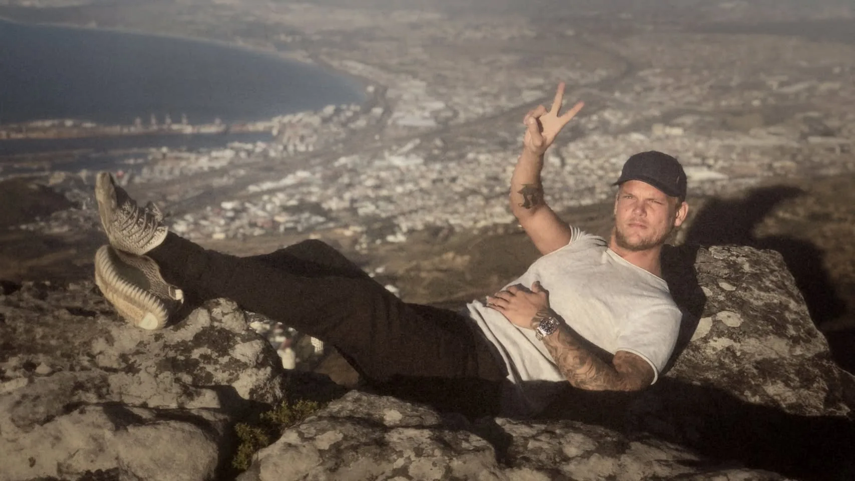 The image depicts a man reclining on a rocky surface, with a scenic view of a coastal city in the background. He is positioned on a large rock, with his legs extended and one arm resting on his stomach. The man is wearing a light gray t-shirt and black pants. His footwear appears to be a pair of patterned sneakers, possibly resembling a popular athletic brand. He has a tattoo visible on his left arm, and he is wearing a watch on his left wrist.

The man is looking directly at the camera with a serious expression, while his right hand is raised in a peace sign. He has short, light-colored hair and is wearing a dark cap. The background features a panoramic view of a city with various buildings and structures, along with a body of water that appears to be a bay or harbor. The landscape is hilly, with some greenery visible in the foreground.

The lighting in the image suggests it may be late afternoon or early evening, casting a warm tone over the scene. The overall atmosphere is relaxed, with the man appearing comfortable in his pose against the natural backdrop. There are no visible logos, text, or signs in the image. The focus remains on the individual and the expansive view behind him.