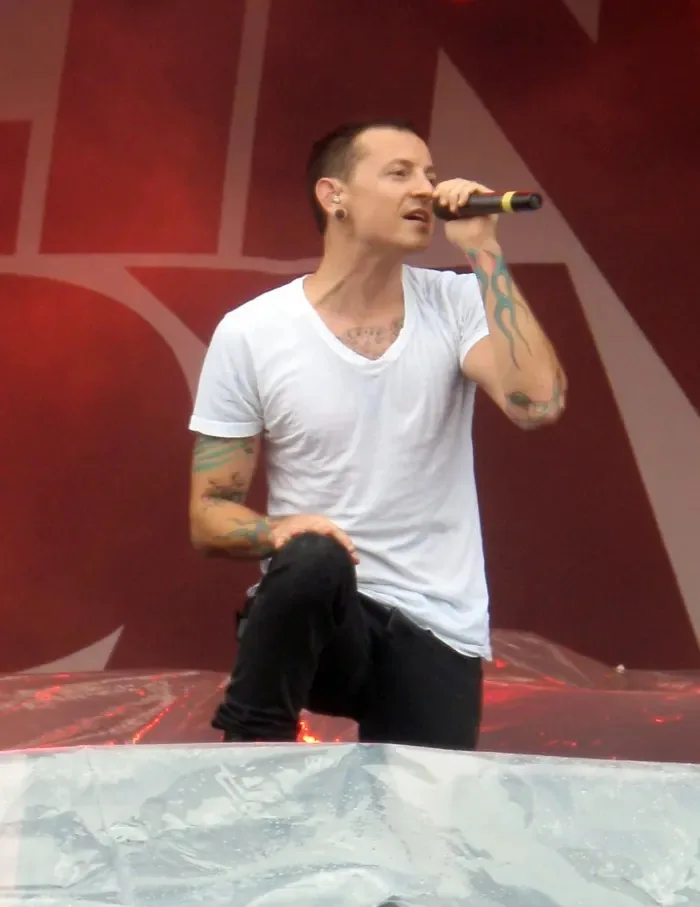 The image depicts a male performer on stage, singing into a microphone. He is wearing a plain white t-shirt that is slightly fitted and has a deep neckline. The shirt appears to be slightly damp, possibly due to rain or sweat. He has black pants that fit snugly, and he is kneeling on a transparent plastic surface, which suggests that the performance may be taking place outdoors, possibly at a festival or concert.

The performer has visible tattoos on both arms, with intricate designs that include colorful patterns. His hair is short and styled, with a slight wave at the front. He has a focused expression on his face, indicating engagement with the performance. The background features a blurred red backdrop with large, bold letters, possibly the name of the band or event, though the text is not clearly readable.

The lighting is dramatic, with a reddish hue dominating the scene, which enhances the atmosphere of the performance. There are no visible logos, brands, or product labels in the image. The overall setting suggests a lively musical event, with an audience likely present, although they are not visible in this particular shot. The focus remains solely on the performer and his dynamic stage presence.