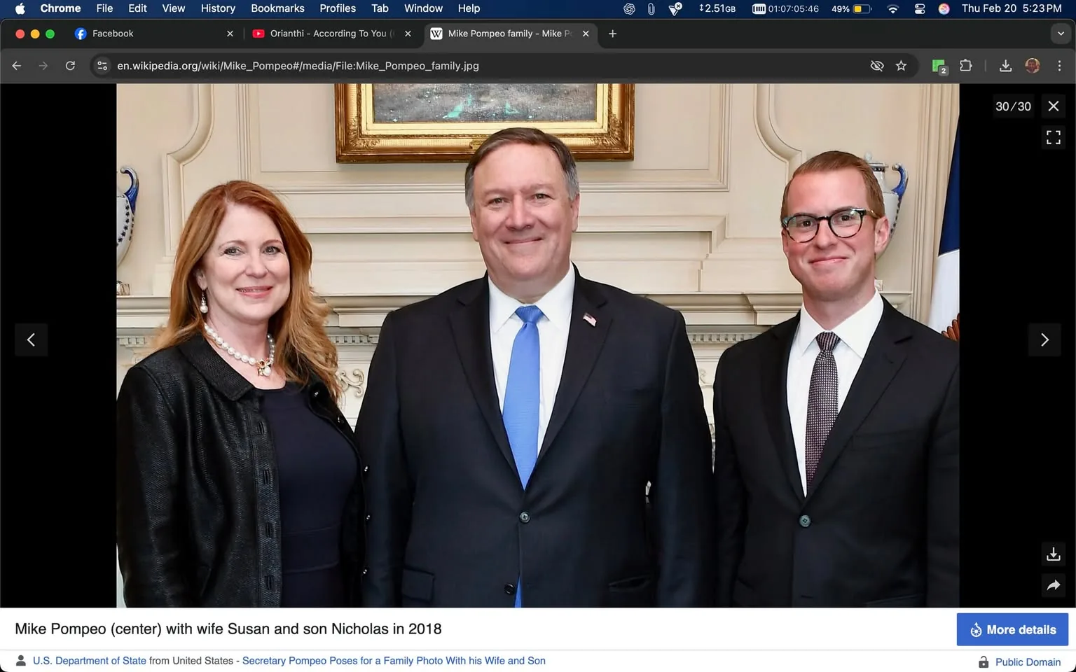 The image depicts three individuals standing together in a formal setting. 

1. **People**:
   - **Left**: A woman with long, wavy reddish-brown hair, wearing a black leather jacket over a dark dress. She has a pearl necklace and is smiling, looking towards the camera.
   - **Center**: A man with short, dark hair, wearing a black suit with a white shirt and a blue tie. He has a small American flag pin on his left lapel and is also smiling, facing the camera directly.
   - **Right**: A young man with short, light brown hair, wearing glasses. He is dressed in a black suit with a white shirt and a patterned tie. He is smiling slightly and looking at the camera.

2. **Clothing**:
   - The woman’s outfit is complemented by her jewelry, which includes a pearl necklace. The leather jacket gives her a polished yet casual appearance.
   - The man in the center is dressed in formal attire, typical for a professional or official event.
   - The young man on the right has a more modern look with his glasses and patterned tie, which adds a touch of style to his formal attire.

3. **Setting**:
   - The background features a light-colored wall with decorative molding. There is a framed painting or artwork above them, which is not clearly visible but adds to the formal ambiance of the setting.
   - To the right, there is a flag, possibly the American flag, indicating a governmental or official context.
   - The overall environment appears to be an indoor space, likely an office or a formal reception area, given the decor and attire of the individuals.

4. **Text**:
   - At the bottom of the image, there is a caption stating: "Mike Pompeo (center) with wife Susan and son Nicholas in 2018." This text identifies the individuals in the photo, suggesting that the man in the center is Mike Pompeo.

5. **Facial Expressions**:
   - All three individuals are smiling, which conveys a sense of positivity and approachability.

Overall, the image captures a formal family portrait in a well-decorated indoor setting, with all subjects dressed in formal attire and displaying friendly expressions.