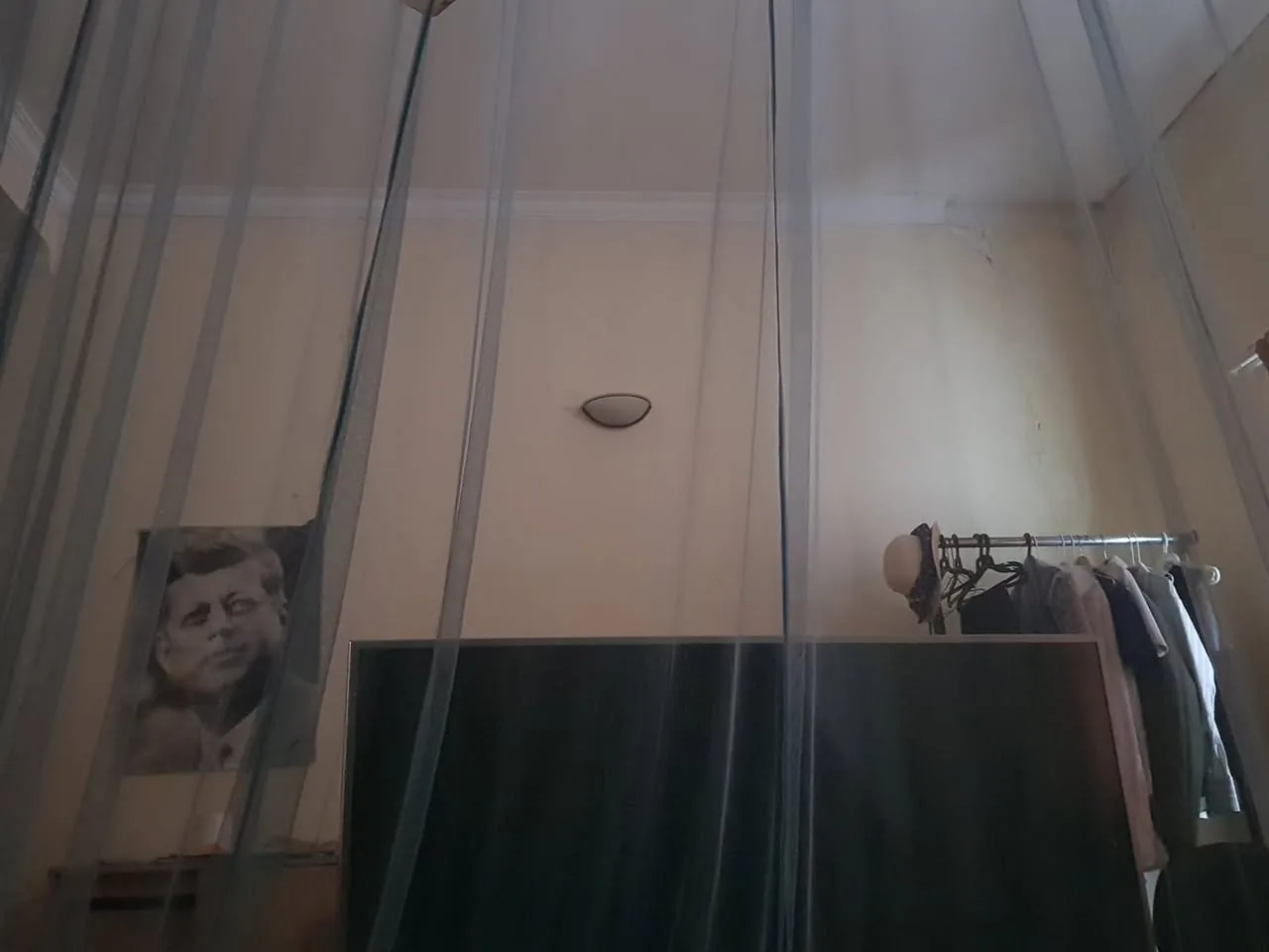 The image depicts an interior space, likely a bedroom, characterized by a soft, muted color palette. The walls are painted a light beige, showing signs of wear with some peeling paint and marks. 

In the foreground, there is a sheer, translucent curtain or netting that hangs from the ceiling, creating a partition or a sense of enclosure within the room. The curtain appears to be made of thin fabric, allowing light to filter through while obscuring the view slightly.

To the left of the image, there is a black-and-white portrait of a man, possibly a public figure, displayed on the wall. The man has a serious expression and is depicted in a classic style, though the details of his features are not clearly visible due to the distance and lighting.

In the center of the image, a large flat-screen television is positioned against the wall. The screen is dark, reflecting the dim lighting of the room. 

To the right, there is a clothing rack with several garments hanging on it. The rack is metallic and holds a variety of clothing items, including shirts and possibly jackets. A hat is also placed on top of the rack, adding to the casual atmosphere of the space.

Above the television, there is a ceiling light fixture, which is round and appears to be turned off, contributing to the overall dim lighting of the room. The ceiling is white and shows some signs of wear, with a few small stains or marks.

Overall, the room has a lived-in feel, with personal items and a cozy atmosphere, but it also shows signs of age and use. The arrangement of objects suggests a functional space, likely used for relaxation or leisure.