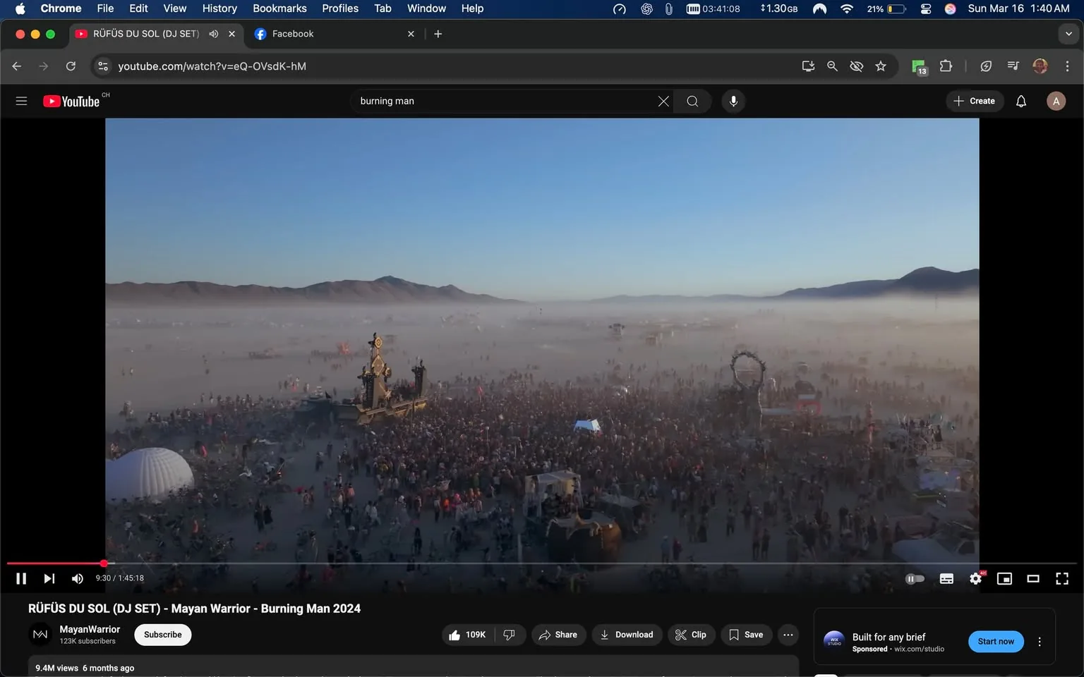 The image depicts a large outdoor gathering at what appears to be the Burning Man festival, characterized by a vast crowd of people in a desert-like environment. The scene is set against a backdrop of mountains, with a clear blue sky above. A significant amount of fog or dust is present, creating a hazy atmosphere.

In the foreground, there is a large crowd of attendees, many of whom are dressed in eclectic and colorful clothing, typical of festival attire. Some individuals are wearing costumes, while others are in casual summer clothing. The crowd appears to be engaged in various activities, with some people standing, others dancing, and a few sitting on the ground.

Prominent in the scene is a large art installation or structure, which is intricately designed and towers over the crowd. It features various elements such as wheels, flags, and possibly lights, indicating it may be part of the festival's artistic displays. To the right, another structure resembling an archway can be seen, adding to the artistic ambiance of the event.

The ground is sandy and dusty, consistent with a desert environment, and there are several tents and temporary structures scattered throughout the area. Some attendees are seen with bicycles, which are commonly used at the festival for transportation.

Overall, the image captures the vibrant and dynamic atmosphere of the Burning Man festival, showcasing the creativity and community spirit of its participants in a unique desert setting.