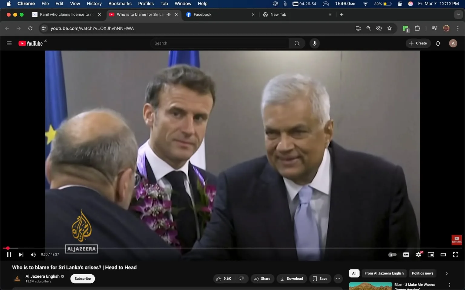 The image appears to be a still from a video featuring three men engaged in conversation. 

1. **People**:
   - **Man on the left**: He has a bald head and is wearing glasses. His expression is not clearly visible as he is turned slightly away from the camera.
   - **Middle man**: He has short, dark hair and is wearing a suit with a white shirt and a black tie. He also has a floral garland around his neck, which includes purple flowers. His expression is serious, and he appears to be looking at the man on the right.
   - **Man on the right**: He has gray hair and is wearing a dark suit with a light blue tie. He is smiling and appears to be extending his hand for a handshake or greeting.

2. **Clothing**:
   - The middle man’s suit is dark, and the floral garland adds a colorful touch. 
   - The man on the right's suit is also dark, with a light blue tie that contrasts with his suit.

3. **Setting**:
   - The background is neutral and appears to be a formal setting, possibly a conference or meeting room. There are flags visible in the background, one of which resembles the European Union flag, indicated by the blue field with yellow stars.

4. **Actions**:
   - The middle man seems to be engaged in conversation with the other two men, while the man on the right is smiling and reaching out, suggesting a friendly interaction.

5. **Text and Logos**:
   - There is a logo in the bottom left corner of the image that reads "Al Jazeera," indicating the source of the video. The video player interface is visible, showing the play button and other controls.

Overall, the image captures a moment of interaction among three men in a formal setting, likely related to a diplomatic or political event.