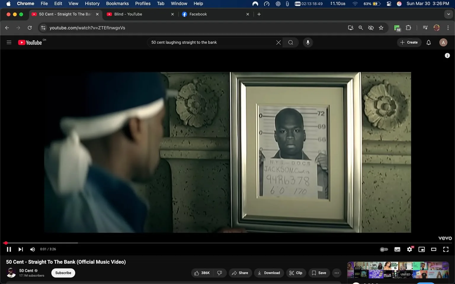 The image depicts a scene from a music video featuring a male figure in the foreground, who is partially turned away from the camera. He is wearing a dark blue outfit and a white bandana tied around his head. The focus of the image is on a framed photograph hanging on the wall behind him. 

The photograph appears to be a police mugshot, featuring a male subject with a shaved head and a neutral expression. The mugshot is labeled with the text "N.Y.S. D.O.C.S. JACKSON, Curtis" along with an identification number "94R6378" and physical details indicating "6'0" and "170" (presumably height and weight). The photograph is framed in a simple, metallic frame and is positioned against a textured wall that has decorative elements, including a floral motif.

The background is dimly lit, creating a somber atmosphere. The wall behind the photograph has a stone-like appearance, with a carved floral design visible to the right of the frame. The overall setting appears to be indoors, possibly in a living room or a similar space, given the decorative nature of the wall and the presence of the framed photograph.

No additional text, logos, or trademarks are visible in the image. The focus remains on the male figure and the mugshot, with no other identifiable brands or objects present. The scene captures a moment of contemplation or reflection, as the male figure gazes at the photograph.