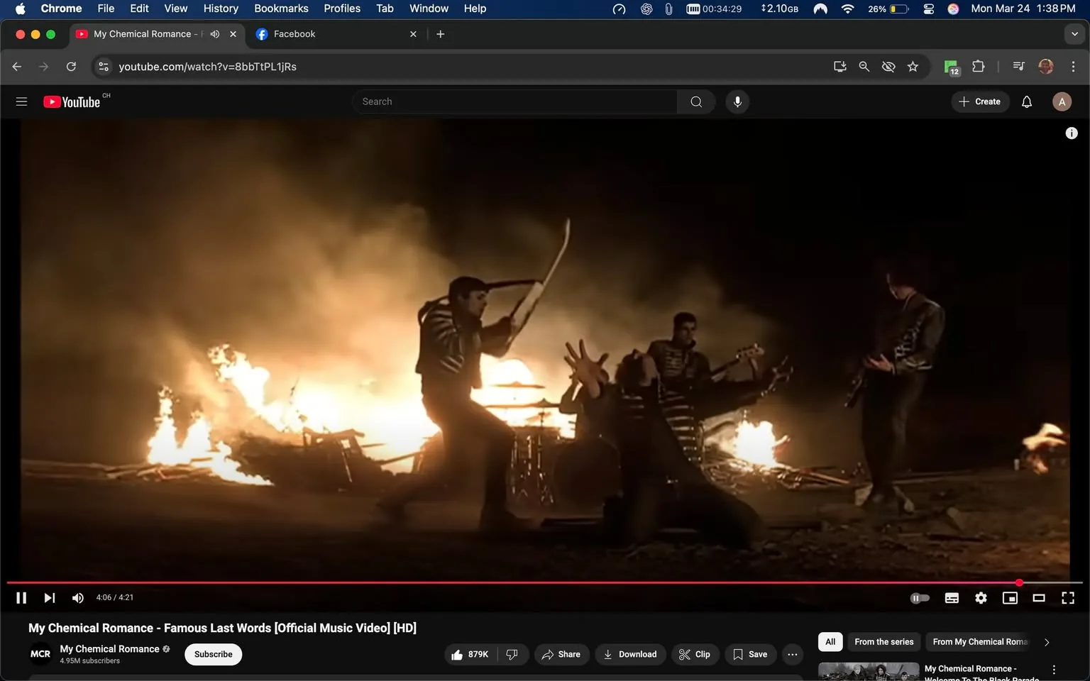 The image depicts a scene from the official music video for "Famous Last Words" by My Chemical Romance. The setting is dark, with a significant fire burning in the background, creating a dramatic and intense atmosphere. The flames illuminate the scene, casting flickering light on the subjects.

In the foreground, there are four individuals, all dressed in black military-style jackets with white stripes. The person on the left is holding a guitar and appears to be in a dynamic pose, possibly playing or performing. Their facial expression is intense, and they have a focused look. The second individual, slightly to the right, is kneeling on the ground with their arms raised, suggesting a dramatic or expressive action. Their face is partially obscured, but they seem to be engaged in the performance.

The third person, positioned further back, is also holding a guitar and appears to be playing. They have a serious expression, and their body language suggests they are fully immersed in the music. The fourth individual, on the far right, is standing with their guitar, looking towards the others. Their posture indicates they are part of the performance.

The background features a large fire, with flames and smoke creating a chaotic yet visually striking backdrop. There are remnants of what appears to be wooden structures or debris near the fire, contributing to the overall dramatic effect of the scene.

The video interface at the bottom includes the YouTube logo, video title "My Chemical Romance - Famous Last Words [Official Music Video] [HD]," and various controls such as play, pause, and volume. The channel name "My Chemical Romance" is displayed, along with a subscriber count of 4.95 million. The video is currently at 4:06 out of a total duration of 4:21.

Overall, the image captures a high-energy moment from a music video characterized by its theatrical presentation and strong visual elements.