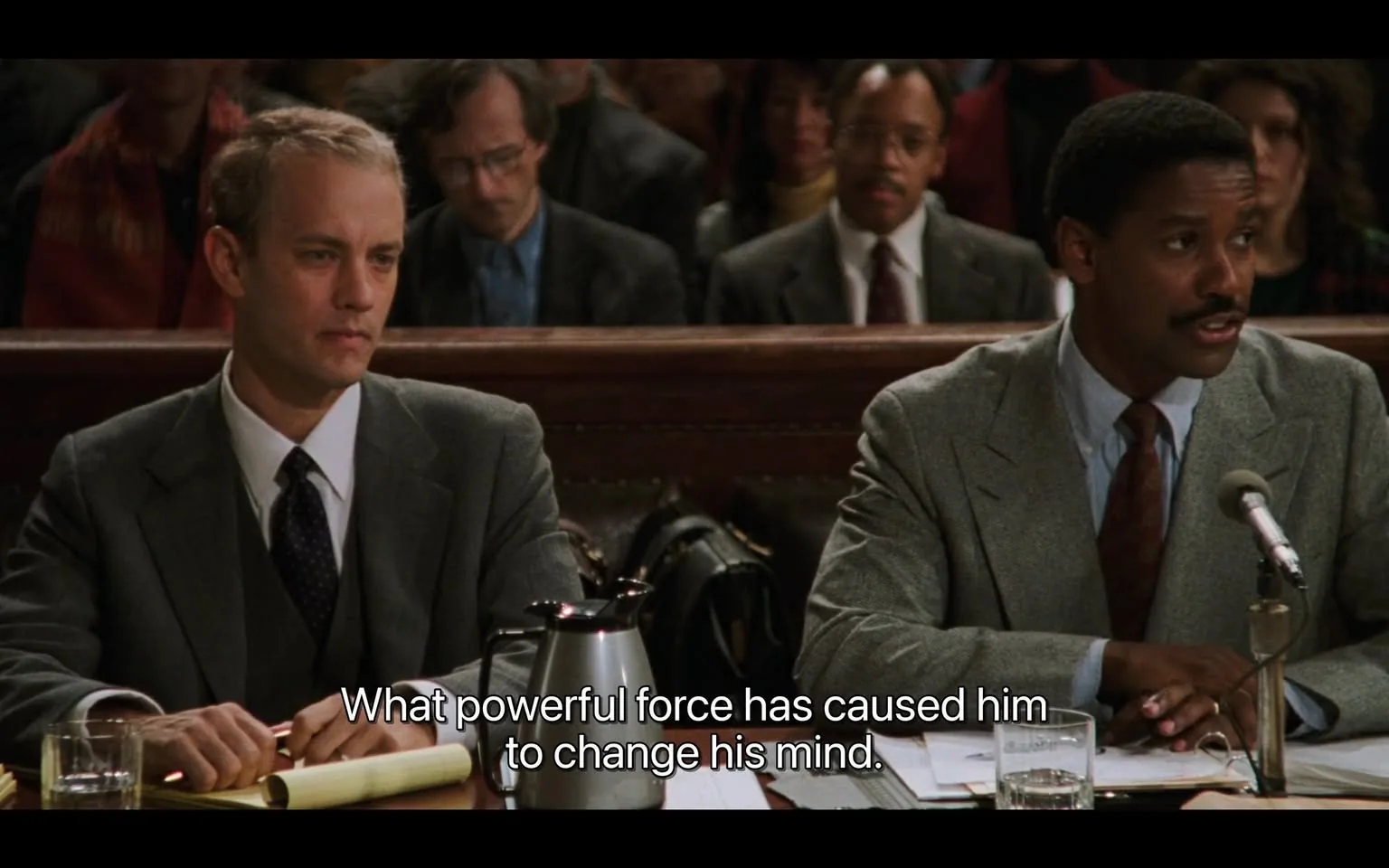 The image depicts a courtroom scene with two men seated at a table in the foreground. 

**Readable Text:**
- There is a line of text at the bottom of the image that reads: "What powerful force has caused him to change his mind."

**People:**
1. **Man on the Left:**
   - He has short, light-colored hair and is wearing a gray suit with a white dress shirt and a dark tie. 
   - His expression appears serious and contemplative as he looks forward, not directly at the camera.
   - He has a glass of water in front of him and is resting his hands on the table.

2. **Man on the Right:**
   - He has dark hair and a mustache, wearing a light gray suit over a white shirt and a patterned tie.
   - He is speaking into a microphone, suggesting he is addressing the court or audience.
   - His expression appears focused and engaged.

**Background:**
- Behind the two men, there are several individuals seated in the courtroom. Some are partially visible, with a mix of expressions and attire. 
- A man in the background is wearing glasses and has curly hair, while another person is wearing a red scarf.
- The setting is a courtroom, indicated by the wooden paneling and the arrangement of seats. The atmosphere appears formal and serious.

**Objects:**
- On the table in front of the two men, there is a black coffee pot, a glass of water, and a yellow legal pad or notebook.
- The table is made of wood and has a polished appearance.

**Overall Setting:**
- The courtroom is dimly lit, with a focus on the two men at the table. The audience in the background is blurred, suggesting they are not the main focus of the image.
- The environment conveys a sense of legal proceedings, with the arrangement of the seating and the presence of a microphone indicating a formal discussion or testimony.

This description captures the observable details in the image without speculation or interpretation.