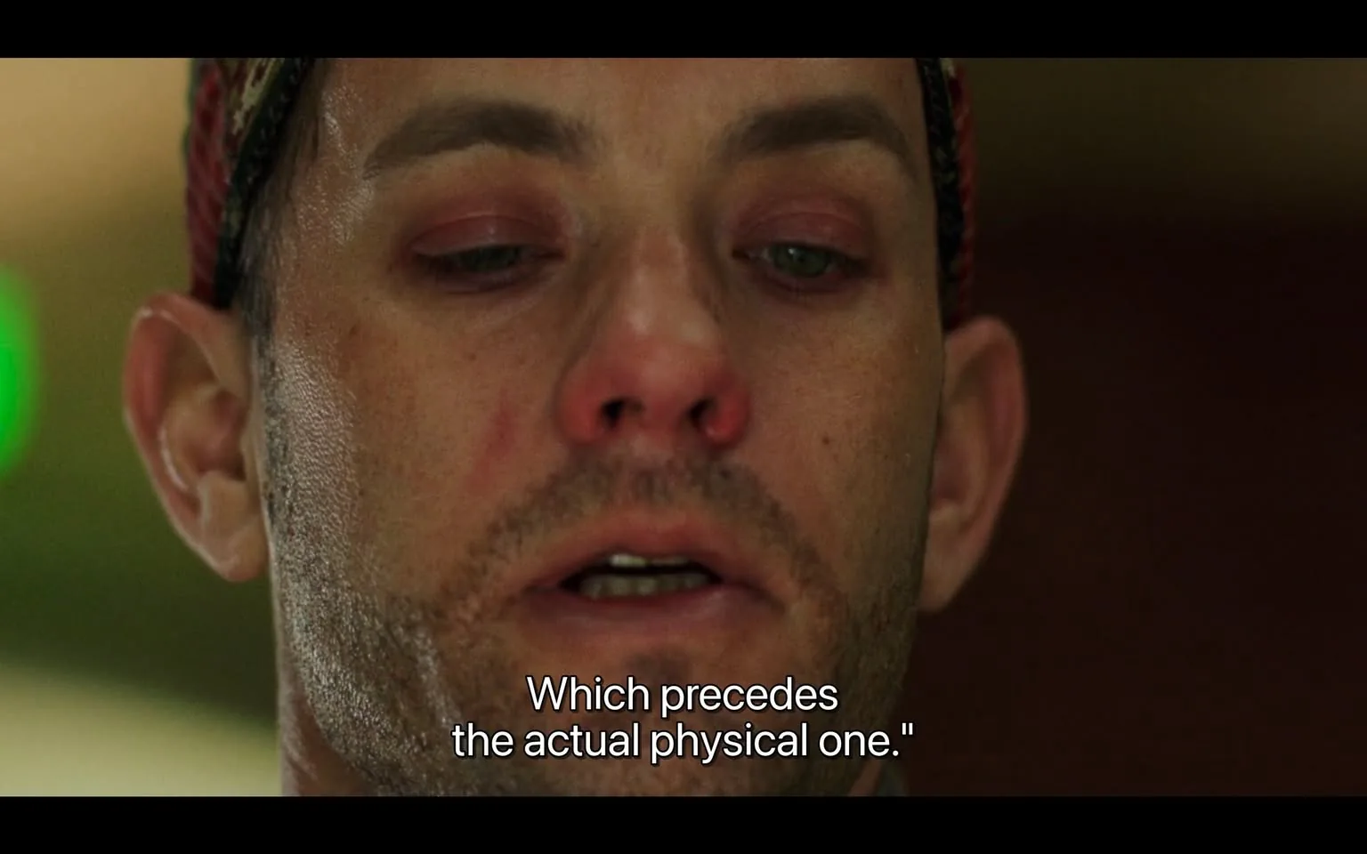 The image features a close-up of a man’s face, capturing a moment of intense expression. His skin appears slightly bruised, with noticeable redness around his nose and under his eyes, suggesting he may have experienced some form of physical altercation or distress. His facial hair is short and stubbly, and he has a light beard. The man has a serious expression, with his mouth slightly open as if he is speaking or about to speak.

His eyes are a striking green, and there is a hint of moisture, possibly indicating emotion or fatigue. The eyelids are slightly swollen, and there is a faint shadow under his eyes, contributing to the overall impression of weariness. He is wearing a patterned headband or beanie, which is primarily red with black and white designs, adding a touch of color to his otherwise neutral-toned appearance.

In the background, there is a blurred green light, which could be a light source or an object, but it is not clearly defined. The setting appears to be indoors, with a soft-focus background that does not provide specific details about the environment. The lighting is warm and somewhat dim, enhancing the mood of the scene.

The text displayed at the bottom reads: "Which precedes the actual physical one." This text is in white font, contrasting against the darker tones of the image, and suggests a dialogue or monologue context, although the source of the quote is not identifiable from the image alone.

Overall, the image conveys a moment of tension, with the man’s physical state and expression drawing attention to his emotional or psychological condition.