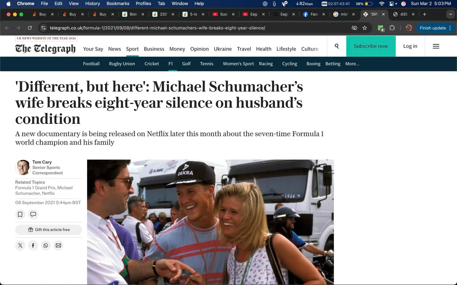 The image is a screenshot of an article from The Telegraph website, featuring a headline that reads: "'Different, but here': Michael Schumacher’s wife breaks eight-year silence on husband’s condition." Below the headline, there is a subheading stating: "A new documentary is being released on Netflix later this month about the seven-time Formula 1 world champion and his family." The article is authored by Tom Cary, who is identified as a Senior Sports Correspondent.

On the left side of the image, there is a photograph showing three individuals. The first person on the left is a man wearing glasses and a black shirt with a white collar, appearing to engage in conversation with the other two. The second person, in the center, is a man with short hair, wearing a black cap with the logo "DEKRA" on it, and a light blue and gray striped shirt. He is smiling and pointing at the woman next to him. The woman, on the right, has long blonde hair and is wearing a white top with a black strap. She is also smiling and appears to be enjoying the interaction.

In the background, there are several large trucks, possibly related to a racing event, and a crowd of people can be seen, indicating a lively atmosphere. The setting appears to be an outdoor event, likely a Formula 1 race, given the context of the article.

The top of the image shows the browser interface, with various tabs open, including social media and other websites. The time displayed is 5:03 PM on a Sunday, March 2, 2021. The overall tone of the article suggests a focus on Michael Schumacher's family and their experiences, particularly in light of a new documentary.