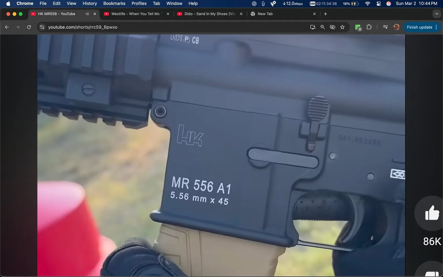 The image displays a close-up view of a firearm, specifically the lower receiver of a rifle. The firearm is marked with the logo "HK," which stands for Heckler & Koch, a well-known firearms manufacturer. The model designation "MR 556 A1" is prominently displayed in white text on the side of the receiver. Below this, the caliber is indicated as "5.56 mm x 45," also in white text. 

The lower receiver features a textured grip area, which is likely designed for better handling. There is a safety selector switch visible, indicating the firearm's operational mode, and a magazine release button is also present. The overall color of the firearm appears to be a matte black, which is common for tactical firearms.

The background is slightly blurred, but hints of green grass and a red object, possibly a container or a piece of equipment, can be seen. The image suggests an outdoor setting, likely a shooting range or training area, given the context of the firearm being displayed.

The hand holding the firearm is gloved, indicating a focus on safety and grip control. The glove appears to be tactical in nature, possibly made of a durable material suitable for handling firearms. The focus of the image is on the firearm itself, with no visible people or additional objects in the immediate foreground. 

Overall, the image captures a detailed view of the firearm's features and markings, set against a natural outdoor backdrop.