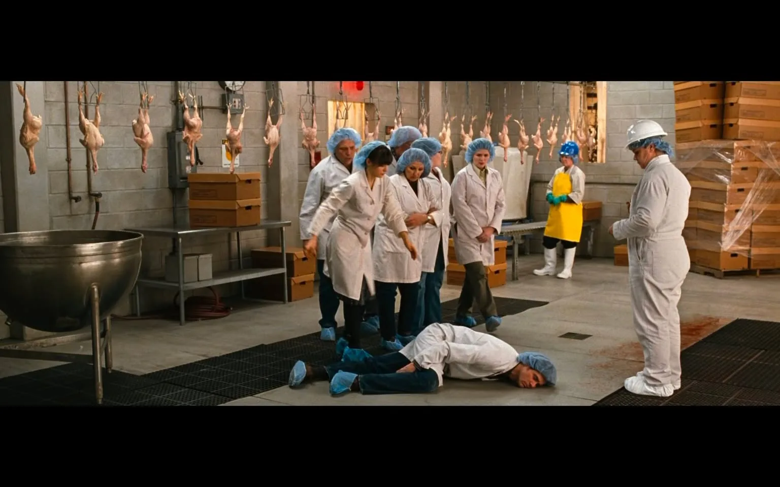 The image depicts a scene inside a meat processing facility. The setting features a large, industrial space with concrete walls and a tiled floor. In the background, several chickens are hanging upside down from hooks, indicating the processing of poultry. There are also large metal containers and cardboard boxes stacked in the area.

In the foreground, a group of people is gathered around a man lying on the floor. He is dressed in a white shirt and blue pants, with blue shoe covers. The man appears to be unconscious or injured, as he is lying face down on the ground. There is a small pool of what appears to be blood near his head, suggesting a serious incident has occurred.

The group surrounding him consists of several individuals wearing white lab coats and blue hairnets. Their expressions are serious, and they seem to be in a state of concern or shock. One woman in the center is reaching out towards the man on the ground, while others are standing close together, observing the situation.

In the background, another person is dressed in a yellow raincoat and white boots, indicating they are also part of the facility's staff. This individual is positioned near a large metal basin, which is empty.

Overall, the scene conveys a sense of urgency and concern, set against the backdrop of a meat processing environment. The lighting is bright, typical of industrial settings, and the overall atmosphere is tense due to the apparent emergency.