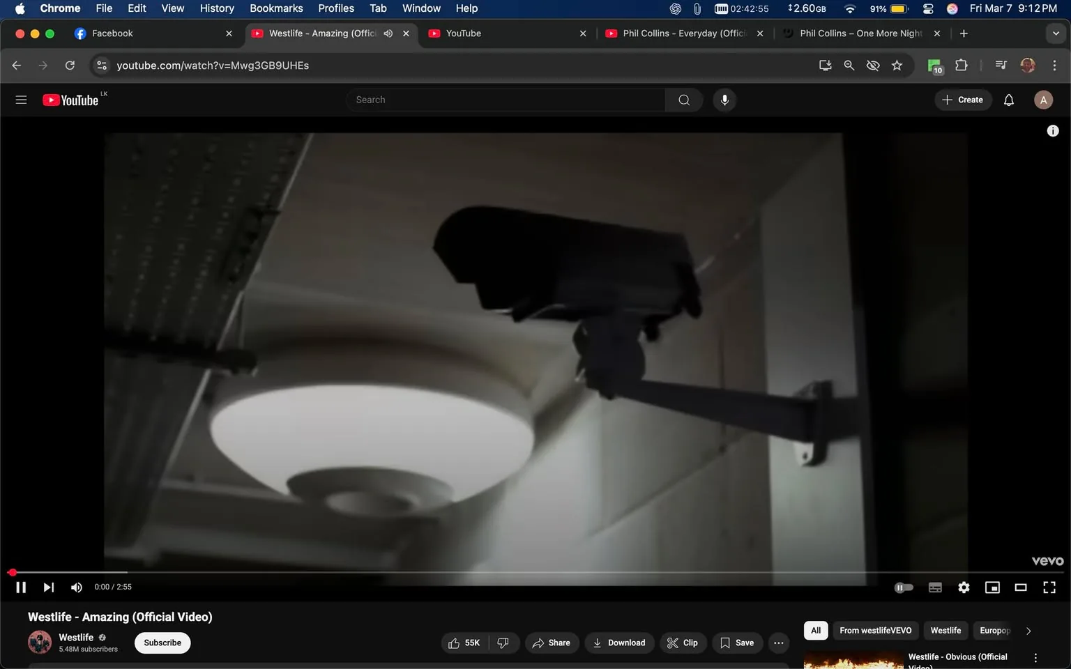 The image shows a surveillance camera mounted on a wall, positioned at an angle that suggests it is monitoring the area. The camera has a black casing with a slightly rounded top and a protruding lens, indicating it is designed for security purposes. It is attached to a white, articulated arm that allows for adjustment in positioning. 

In the background, there is a ceiling light fixture that emits a soft, diffused light, creating a warm ambiance in the space. The light fixture has a circular design with a central bulb surrounded by a circular cover, which is likely made of plastic or glass. The ceiling appears to be a light color, possibly white or off-white, and has a smooth finish.

The walls adjacent to the camera are also light-colored, and there are visible seams or joints that suggest a paneling or drywall construction. The overall setting appears to be indoors, possibly in a hallway or a room that requires monitoring for security reasons.

There are no visible people, text, or logos in the image. The focus is solely on the surveillance camera and the surrounding environment. The lighting and colors suggest a neutral, unobtrusive atmosphere, typical of spaces where surveillance is necessary.