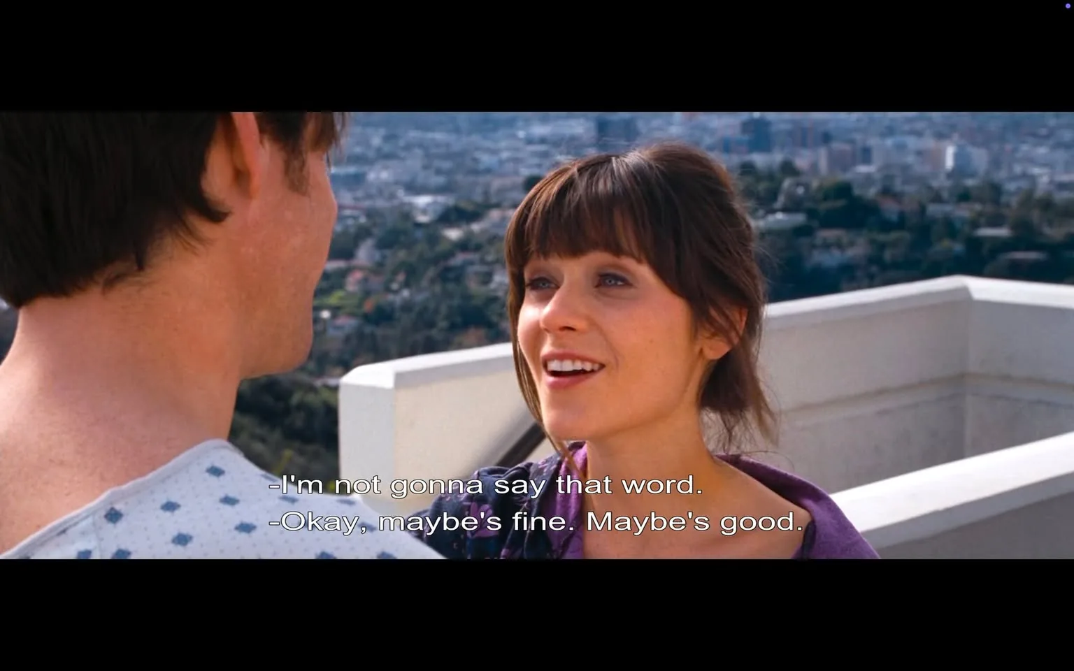 The image depicts a scene from a film featuring two characters engaged in conversation. The character on the left is a man, whose back is turned towards the camera. He has short, dark hair and is wearing a hospital gown with a blue and white pattern, which suggests a medical setting. His skin tone appears fair, and he has a slight stubble on his face.

The character on the right is a woman with brown hair styled in soft waves and bangs. She is facing the camera and smiling, displaying a friendly and animated expression. She is wearing a purple top, which contrasts with the lighter background. The woman has a light complexion and is looking directly at the man, indicating engagement in their conversation.

In the background, there is a panoramic view of a cityscape, with numerous buildings and greenery visible, suggesting a high vantage point. The sky is clear, and the setting appears to be outdoors, possibly on a rooftop or balcony.

The dialogue displayed at the bottom of the image reads:
- "I'm not gonna say that word."
- "Okay, maybe's fine. Maybe's good."

This text indicates a light-hearted exchange between the characters. The overall atmosphere of the scene appears casual and intimate, with the characters in a moment of personal interaction. The background adds depth to the setting, enhancing the visual context of the conversation.