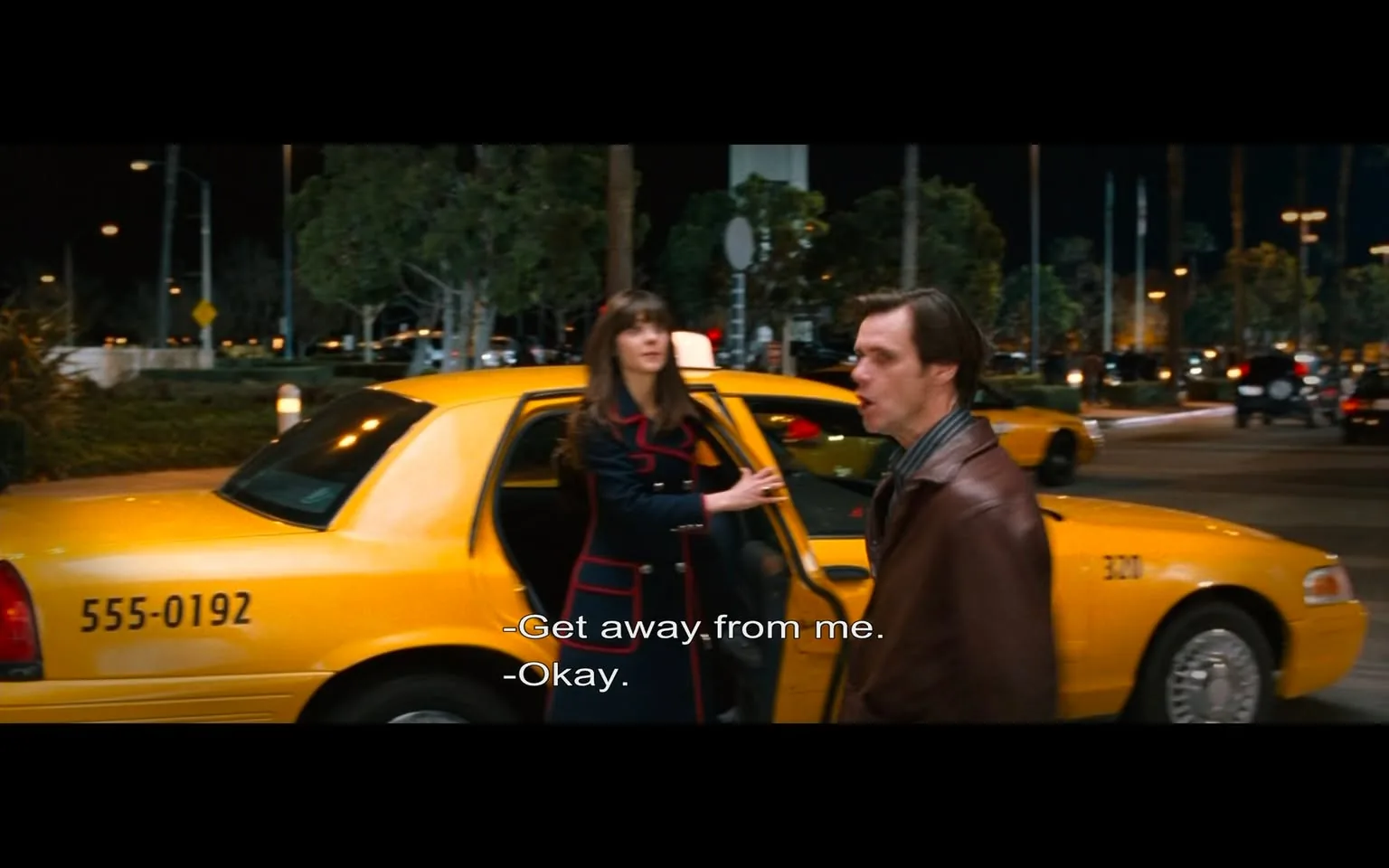 The image depicts a scene set at night, featuring a yellow taxi cab parked on a street. The taxi has a visible number on its side: "555-0192." The background shows a well-lit area with trees and other vehicles, indicating a busy urban environment. 

In the foreground, there are two individuals engaged in a conversation. On the left, a woman is standing next to the open door of the taxi. She has long, dark hair and is wearing a dark coat with red accents. Her expression appears to convey frustration or assertiveness as she gestures with her hand, saying, "Get away from me."

On the right, a man is standing slightly turned away from her. He has short, light brown hair and is wearing a brown leather jacket over a striped shirt. His facial expression seems to reflect surprise or concern as he responds with, "Okay." 

The lighting in the scene is bright, likely from streetlights and the taxi's interior light, creating a contrast with the darker surroundings. The setting suggests a public space, possibly near an airport or a busy street, given the presence of other cars and the taxi. The overall atmosphere appears tense, as indicated by the body language and expressions of the two individuals.