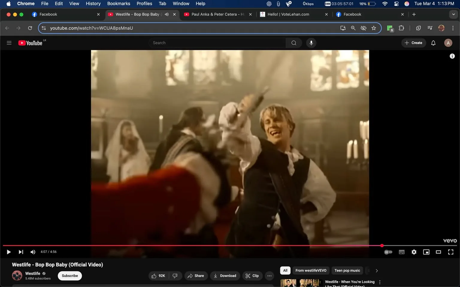 The image shows a scene from a music video titled "Bop Bop Baby" by Westlife. The setting appears to be an ornate interior, likely a church or a grand hall, characterized by high ceilings and large stained glass windows that allow soft, diffused light to filter through. The atmosphere is somewhat dramatic, with a warm color palette dominated by browns and golds.

In the foreground, a male figure is prominently featured, smiling and engaging in a lively action. He has short, light-colored hair and is dressed in a historical-style outfit, which includes a white shirt with billowy sleeves and a dark vest. His expression is joyful, and he appears to be playfully wielding a sword, which is pointed upward. The sword has a shiny blade and a decorative hilt. 

Behind him, there are additional figures dressed in similar historical attire, suggesting a choreographed dance or performance. One figure, partially visible, appears to be wearing a long, flowing gown and a veil, indicating a possible wedding theme. The background includes tall candlesticks with lit candles, enhancing the ambiance of the scene.

The video interface at the bottom of the image displays the YouTube logo and various controls, including play, pause, and volume settings. The title "Westlife - Bop Bop Baby (Official Video)" is visible, along with the channel name "Westlife" and subscriber count of 5.48 million. The video is currently at 4:07 out of a total duration of 4:56.

Overall, the image captures a moment of energetic performance within a richly detailed historical setting, emphasizing the playful and upbeat nature of the song.