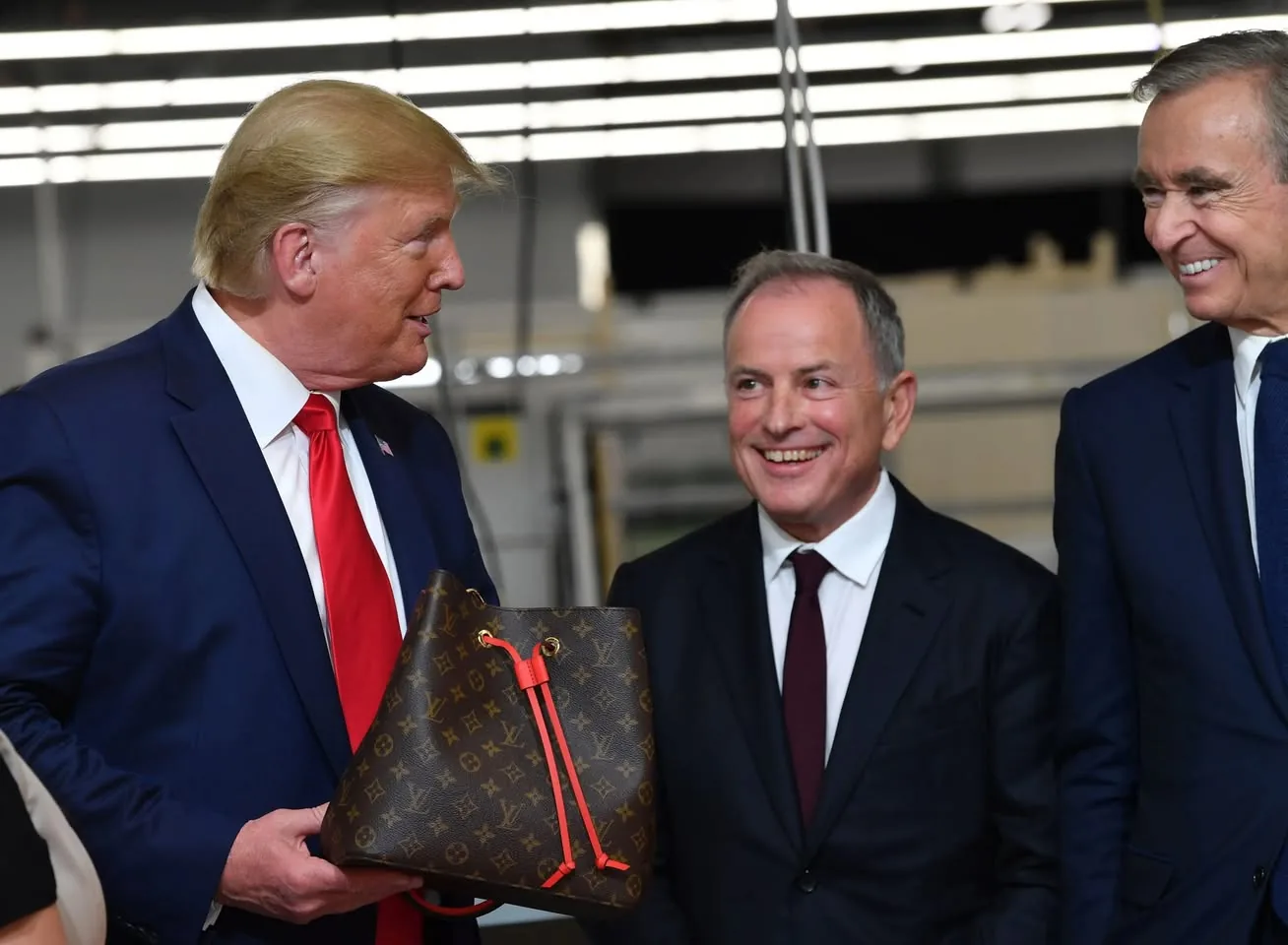 The image depicts three men engaged in conversation, with one man holding a handbag. 

1. **People**:
   - The man on the left has light blonde hair styled back and is wearing a dark suit with a white shirt and a red tie. He is smiling and appears to be speaking, looking towards the man on his right.
   - The man in the center has short, dark hair with some gray, wearing a dark suit with a white shirt and a maroon tie. He is smiling broadly and looking at the man on the left.
   - The man on the right has dark hair and is also wearing a dark suit with a white shirt. He is smiling and looking at the man on the left, appearing engaged in the conversation.

2. **Handbag**:
   - The handbag being held by the man on the left is a brown Louis Vuitton bag, featuring the brand's signature monogram pattern. It has a bright red drawstring and accents, which stand out against the brown background.

3. **Setting**:
   - The background appears to be a well-lit indoor space, possibly a factory or showroom, with overhead fluorescent lights. There are blurred outlines of machinery or shelving in the background, indicating a commercial or industrial environment.

4. **Facial Expressions**:
   - All three men are smiling, suggesting a positive interaction. The expressions convey engagement and friendliness.

5. **Clothing**:
   - The clothing of the men is formal, consisting of suits and ties, which is typical for a business or formal event.

Overall, the image captures a moment of interaction among three men, with a focus on the handbag being presented. The setting and attire suggest a professional context.