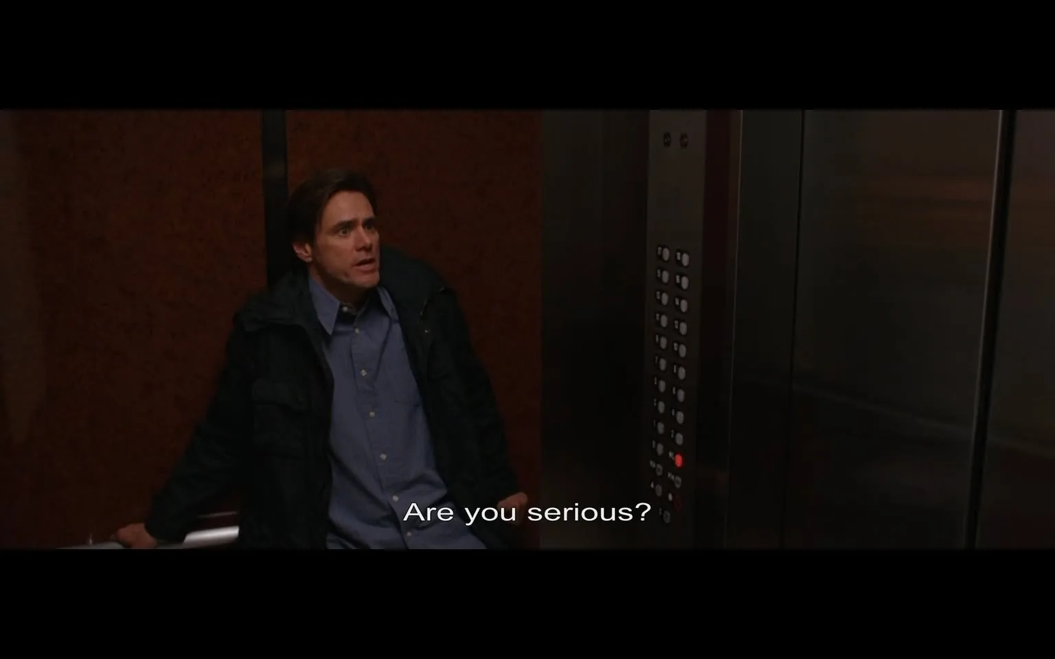 The image depicts a scene inside an elevator. The walls of the elevator are a reddish-brown color, possibly a textured material, giving a warm but somewhat dated appearance. The elevator doors are metallic and reflect some light, indicating a polished surface.

In the foreground, a man is seated against the wall of the elevator. He has short, dark hair and is wearing a black jacket over a blue button-up shirt. His expression appears to convey surprise or disbelief, as he looks slightly upward with his mouth slightly open, suggesting he is in the midst of speaking. The text overlay reads, "Are you serious?" indicating a moment of dialogue.

To the right of the man is the elevator control panel, which features a series of buttons. The buttons are arranged in a grid format, with some illuminated, indicating the current floor or selection. One button is lit up in red, possibly indicating the floor the elevator is currently on or a call button.

The overall setting is confined and enclosed, typical of an elevator environment, with no visible windows or external views. The lighting is soft, likely artificial, contributing to the intimate atmosphere of the scene. There are no other people visible in the image, and the focus remains solely on the man and the elevator's interior.