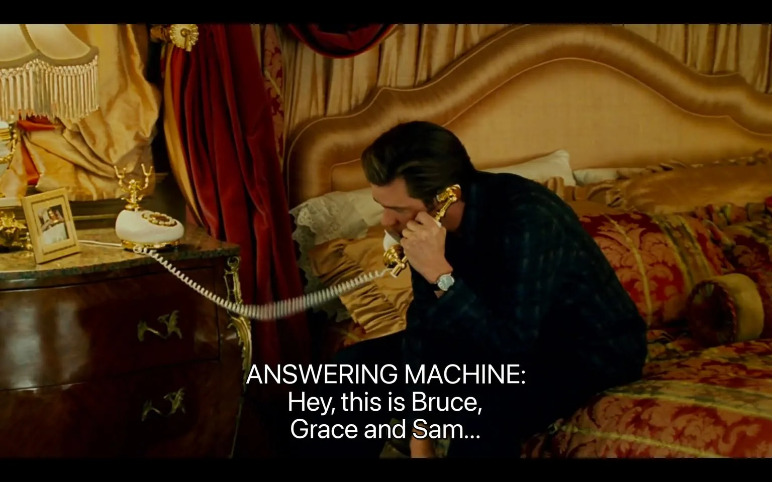 The image depicts a scene in a bedroom setting. A man is seated on a bed, holding a golden rotary telephone to his ear. He appears to be engaged in a conversation. The man has dark hair styled back and is wearing a dark plaid shirt. His facial expression is focused, suggesting he is listening intently.

The bed is prominently featured, with a large, upholstered headboard in a light color. The bedding is richly textured, with a mix of gold and red tones, and there are decorative pillows scattered across the bed. A bedside table is visible next to the bed, made of dark wood, with ornate detailing. 

On the table, there is a white rotary telephone, which has a coiled cord connecting it to the base. Next to the phone, there is a small picture frame containing a photo, though the contents of the photo are not discernible. The frame is positioned in front of a decorative object that resembles a small golden antler or branch.

In the background, heavy drapes in a rich red color frame a window, adding to the opulent feel of the room. The lighting appears warm and soft, likely from a lamp with fringe detailing, which is also visible in the scene. 

The text displayed on the image reads: "ANSWERING MACHINE: Hey, this is Bruce, Grace and Sam..." This text is positioned at the bottom of the image in a bold, white font, contrasting against the darker background. 

Overall, the setting conveys a luxurious and intimate atmosphere, characterized by rich colors and textures, with the man engaged in a private conversation.