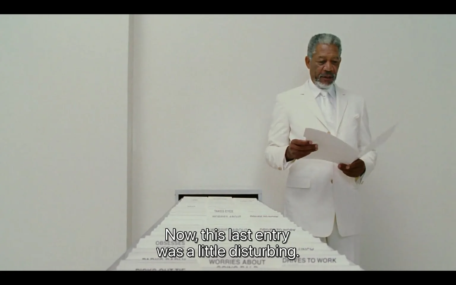 The image features a man standing in a minimalist, brightly lit space with a plain white background. He is dressed in a formal white suit, which includes a jacket, trousers, and a dress shirt, complemented by a white tie. The suit appears well-tailored, and he has a neatly groomed beard and hair, which is gray with some darker strands. His expression is serious as he holds a few sheets of paper in his right hand and appears to be reading from them.

In front of him is a long table or surface that is lined with numerous sheets of paper, each labeled with different phrases. The labels visible on the papers include "TAKES EYES," "WORRIES ABOUT," "OBSESSES," and "DRIVES TO WORK." The papers are arranged in a neat, organized manner, suggesting a systematic approach to the information presented. The table is devoid of any additional objects or decorations, emphasizing the simplicity of the setting.

The overall environment is stark and clinical, with no visible furniture or decor aside from the table and the papers. The lighting is bright and even, contributing to the clean aesthetic of the space. The man’s posture and the way he holds the papers indicate that he is engaged in a presentation or explanation of the contents. There are no identifiable logos, brands, or trademarks present in the image.