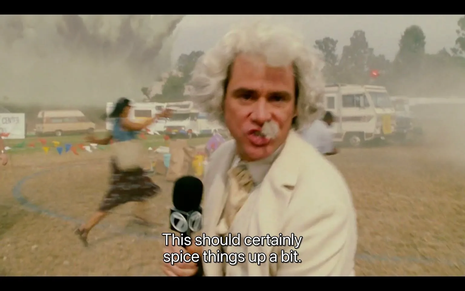 The image depicts a chaotic scene with a man in the foreground, who appears to be a news reporter. He has a distinctive hairstyle characterized by curly, white hair and a mustache. He is dressed in a light-colored suit, which is slightly wrinkled, and he is holding a microphone with a logo that resembles a news channel emblem, possibly indicating a television network. The text on the screen reads, "This should certainly spice things up a bit," suggesting he is reporting on an unfolding event.

In the background, there is a significant weather phenomenon, likely a tornado or a large storm cloud, creating a dramatic and tense atmosphere. The sky is dark and ominous, with swirling clouds that suggest turbulence. 

To the left of the reporter, a woman is seen running away from the camera, wearing a blue top and a long skirt. Her facial expression appears to convey urgency or fear as she points in the direction of the storm. There are also several people in the background, some of whom seem to be reacting to the situation, with a few others visible in the distance near parked vehicles, including RVs and trailers. 

Colorful triangular flags are strung across the scene, indicating that this might have been a festive event, possibly a fair or gathering, before the storm disrupted it. The ground is covered in dirt and appears to be a field or open area. 

Overall, the image captures a moment of tension and urgency, with the reporter in focus amidst a backdrop of chaos and environmental disturbance.