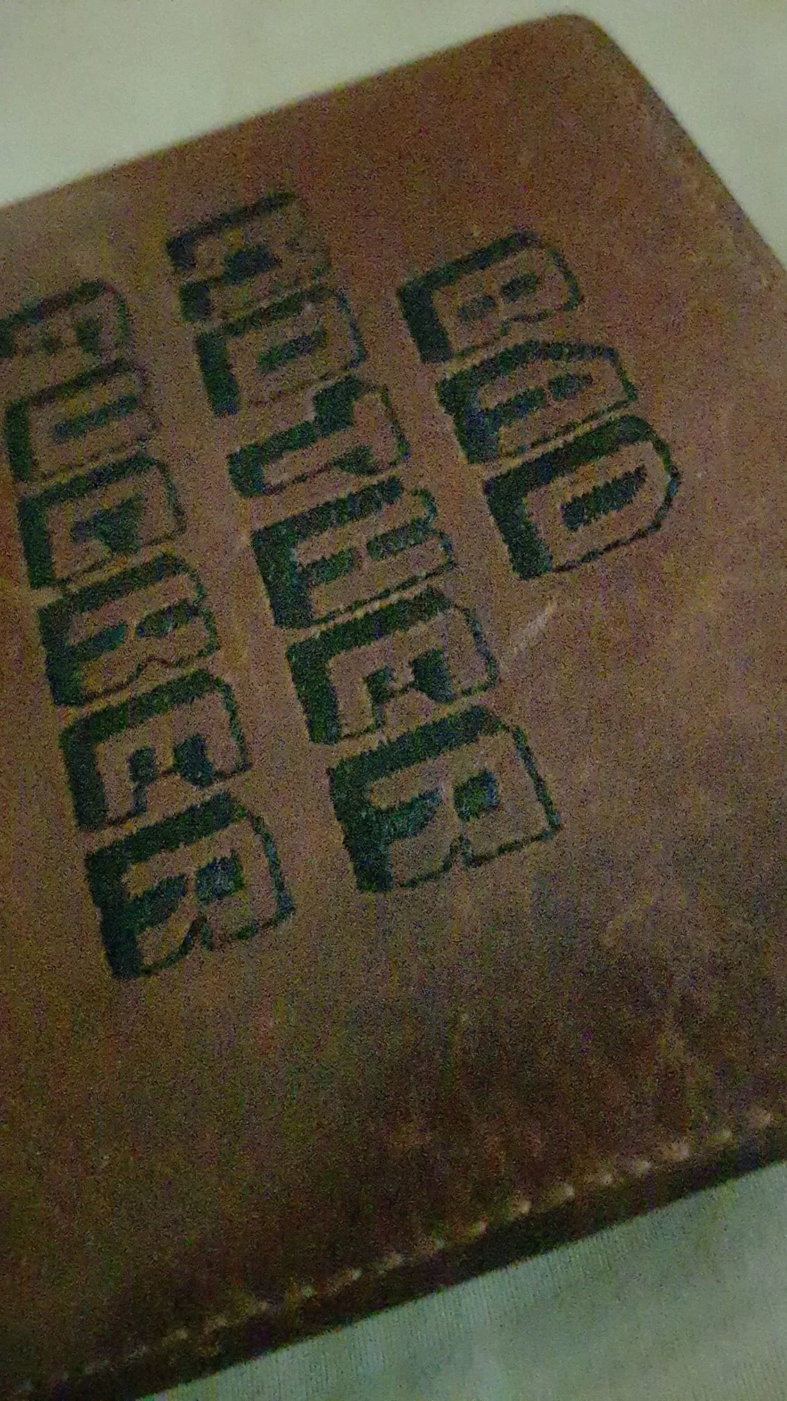 The image features a close-up view of a brown leather wallet or similar item. The wallet is partially open, revealing a textured surface. Prominently displayed on the front cover is the text "BAD MOTHER F***ER," which is printed in bold, black, block letters. The lettering appears slightly distressed, suggesting wear or age, with some areas showing a faded quality. The text is centered and occupies a significant portion of the wallet's surface.

The wallet itself has a smooth finish, with visible stitching along the edges, indicating craftsmanship. The background is a light-colored surface, possibly a table or cloth, which contrasts with the darker tones of the wallet. The lighting in the image is somewhat dim, contributing to a slightly grainy appearance, but the text remains legible.

No people are present in the image, and there are no additional objects or items visible in the frame. The setting appears to be indoors, but no specific location or environmental context is provided. There are no logos, brands, or trademarks visible aside from the text on the wallet. The overall focus is on the wallet and its distinctive text.