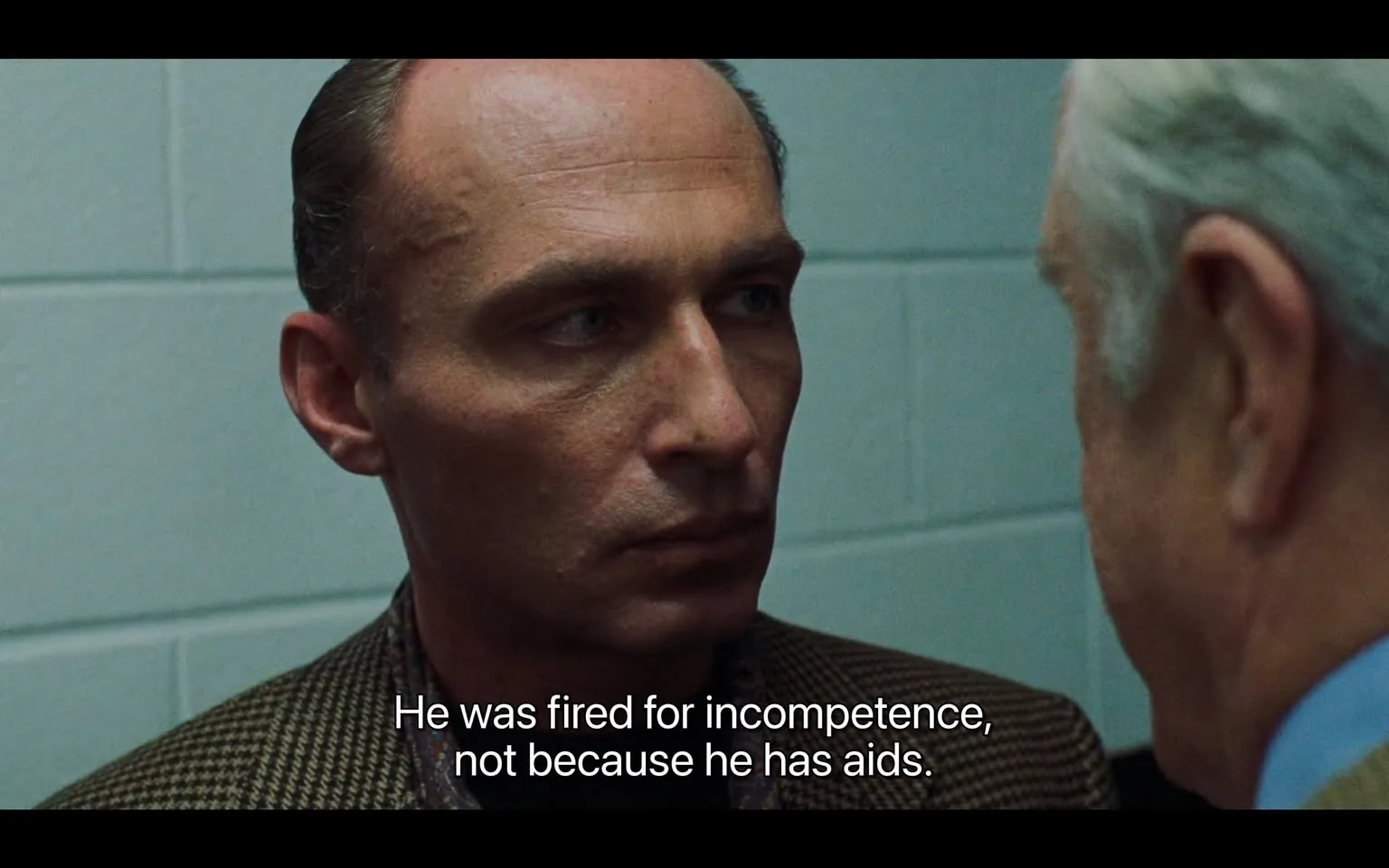 The image depicts a close-up scene featuring two men engaged in a serious conversation. 

**Readable Text:**
- There is a line of dialogue displayed at the bottom of the image: "He was fired for incompetence, not because he has aids."

**People:**
1. **Man on the Left:**
   - He has a shaved head and a light complexion.
   - His facial expression appears serious and intense, with a slight furrow in his brow.
   - He is wearing a brown checkered suit jacket over a white shirt, which is partially visible.
   - His gaze is directed towards the man on the right, indicating engagement in the conversation.

2. **Man on the Right:**
   - He has white hair and a light complexion.
   - His facial expression is also serious, and he appears to be speaking or responding to the man on the left.
   - He is wearing a light-colored suit, possibly beige or gray, with a white shirt underneath.
   - His gaze is directed towards the man on the left, suggesting he is listening intently.

**Setting:**
- The background consists of a light blue wall, which appears to be made of cinder blocks, suggesting an indoor environment, possibly an office or a meeting room.
- The lighting is somewhat dim, contributing to the serious tone of the scene.

**Overall Composition:**
- The focus is primarily on the two men, with their faces taking up a significant portion of the frame.
- The image captures a moment of tension or confrontation, as indicated by their expressions and proximity to each other.

This description captures the observable details in the image without speculation or interpretation.