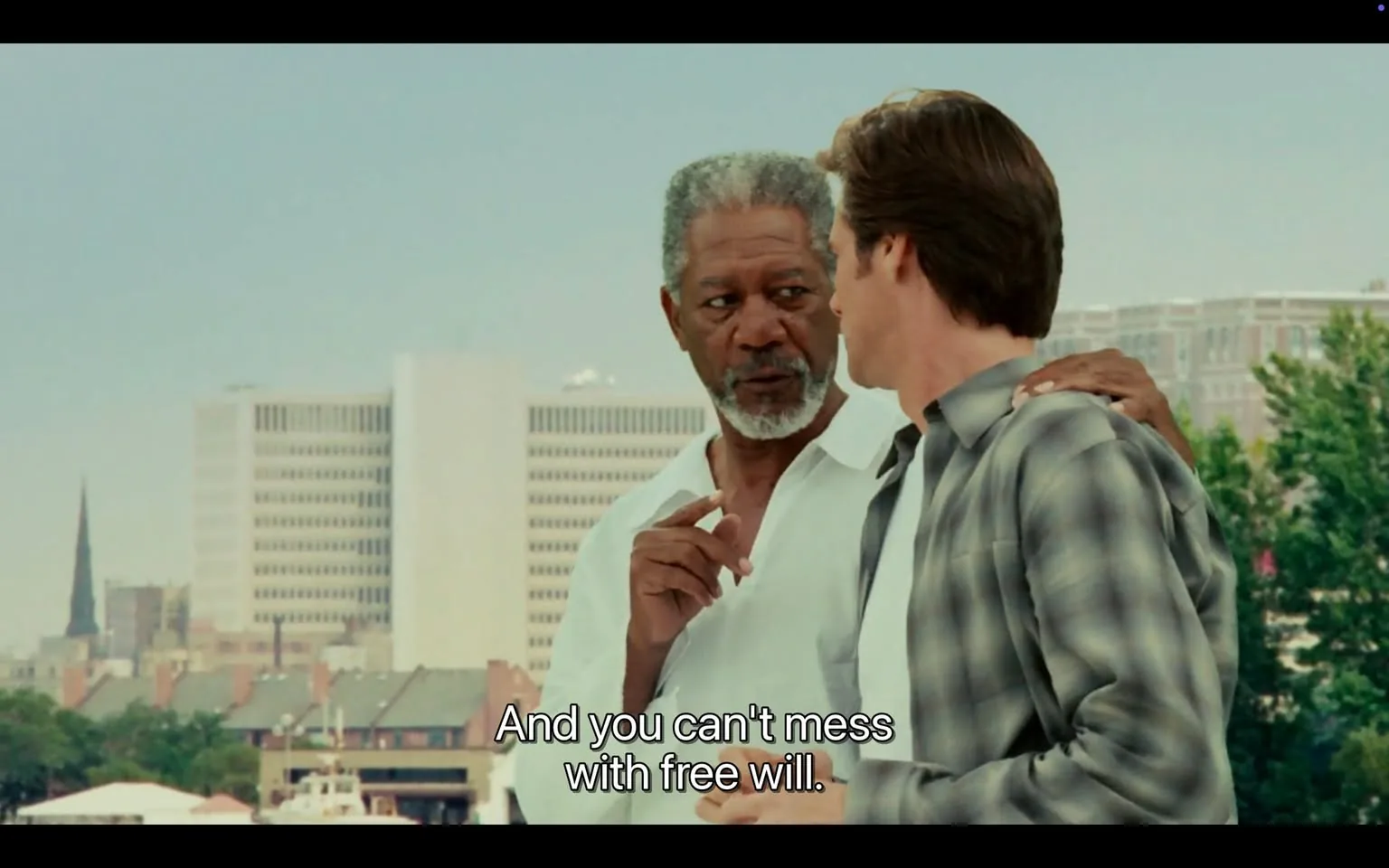 The image features two men engaged in conversation against a backdrop of a cityscape. 

**Readable Text:**
At the bottom of the image, there is a subtitle that reads: "And you can't mess with free will."

**People:**
1. **First Man:**
   - **Appearance:** He has gray hair and a beard, which is also gray. His skin tone is darker.
   - **Clothing:** He is wearing a white shirt, which appears to be buttoned down.
   - **Facial Expression:** He has a serious expression and is looking intently at the other man.
   - **Action:** He is gesturing with his right hand, possibly emphasizing a point in the conversation.

2. **Second Man:**
   - **Appearance:** He has brown hair and a lighter skin tone.
   - **Clothing:** He is wearing a gray and black plaid shirt over a white undershirt.
   - **Facial Expression:** He appears to be listening attentively to the first man.
   - **Action:** He is standing close to the first man, with his left hand resting on the first man's shoulder.

**Setting:**
- The background features a city skyline with several buildings. There is a prominent structure that appears to be a high-rise building with a grid-like facade.
- In the distance, there is a spire of a church or similar structure, indicating a mix of modern and possibly historical architecture.
- The foreground shows rooftops of buildings, suggesting an urban environment. There are trees visible, indicating greenery in the area.

**Environmental Context:**
- The scene appears to be outdoors, likely on a rooftop or elevated area, given the perspective of the skyline.
- The lighting suggests it is daytime, with a clear sky and no visible clouds.

Overall, the image captures a moment of dialogue between two men in a city setting, with a focus on their expressions and gestures.