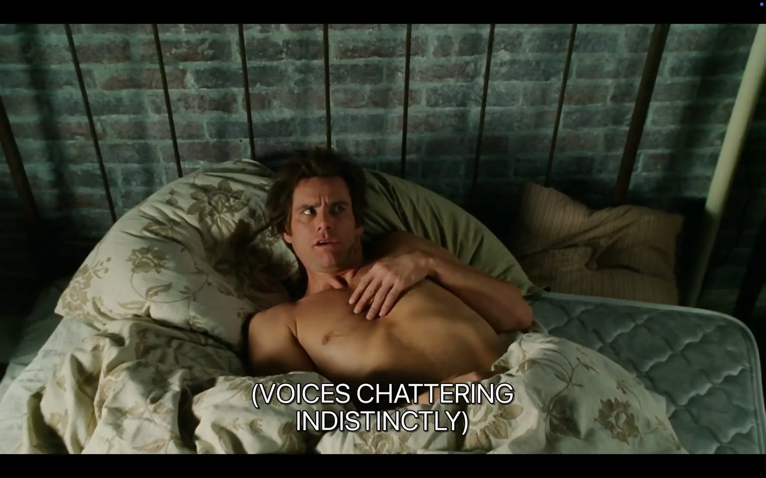 The image depicts a man lying in bed, appearing startled or confused. He has long, slightly tousled hair and is shirtless, with his upper body visible. His expression shows a mix of surprise and concern, with wide eyes and slightly raised eyebrows. He is positioned on a bed with a patterned comforter that features floral designs in muted colors. The bedding appears to be slightly rumpled, suggesting he has just woken up or is in a state of disarray.

The background consists of a brick wall, which is painted in a light color, giving a rustic feel to the setting. There are two pillows visible at the head of the bed, one of which is slightly crumpled. The overall lighting in the scene is soft, indicating it might be early morning or late evening.

At the bottom of the image, there is a subtitle that reads "(VOICES CHATTERING INDISTINCTLY)," suggesting that there is audio content accompanying the visual, likely indicating a scene from a film or television show. 

No logos, brands, or identifiable trademarks are present in the image. The setting appears to be a bedroom, but no additional context about the location or event is provided. The focus remains on the individual and his immediate surroundings.