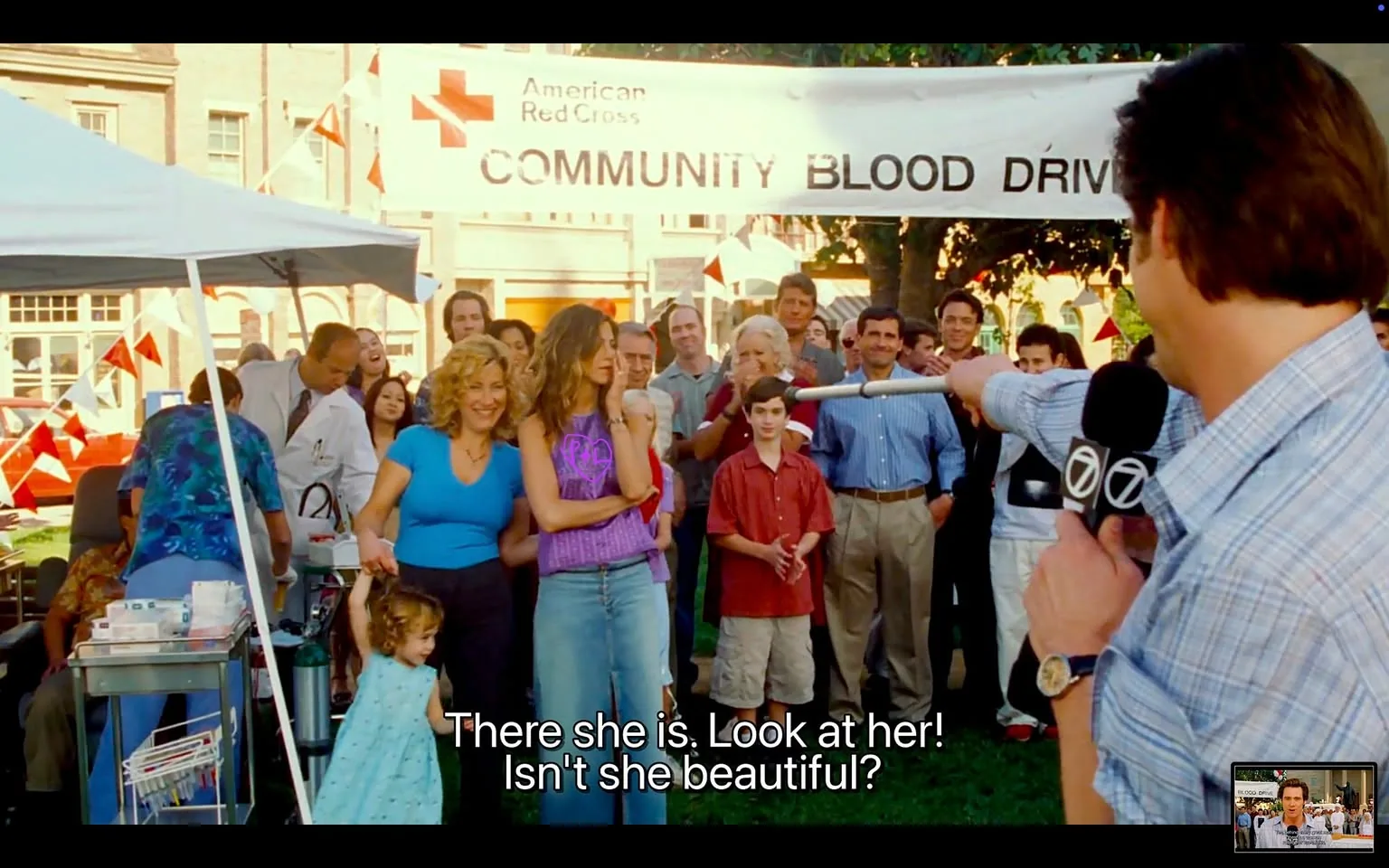 The image depicts a community event, specifically a blood drive organized by the American Red Cross. A large banner hangs prominently in the background, reading "COMMUNITY BLOOD DRIVE" with the American Red Cross logo visible. The setting appears to be an outdoor area, likely a park or community square, with several people gathered around.

In the foreground, a man holding a microphone is addressing the crowd. He is wearing a light blue checkered shirt and appears to be pointing towards a woman in the crowd. The man has short, dark hair and is looking towards the audience with a smile. The crowd consists of a diverse group of people, including men, women, and children, all engaged and looking towards the speaker.

The woman being pointed at is in the center of the image, wearing a purple sleeveless top and light blue jeans. She has long, wavy hair and is standing with her arms crossed, displaying a neutral expression. To her left, a woman with curly hair is smiling and wearing a turquoise top. A young girl in a light blue dress is holding the woman's hand. 

In the background, various individuals can be seen, including a boy in a red shirt and another man in a blue striped shirt. There are also several adults in business attire, suggesting a mix of community members and possibly local officials. The environment is decorated with red and white banners, and there are tables set up, likely for the blood donation process, with medical equipment visible.

Overall, the scene conveys a lively community atmosphere, focused on the blood drive event, with a mix of excitement and engagement from the attendees.
