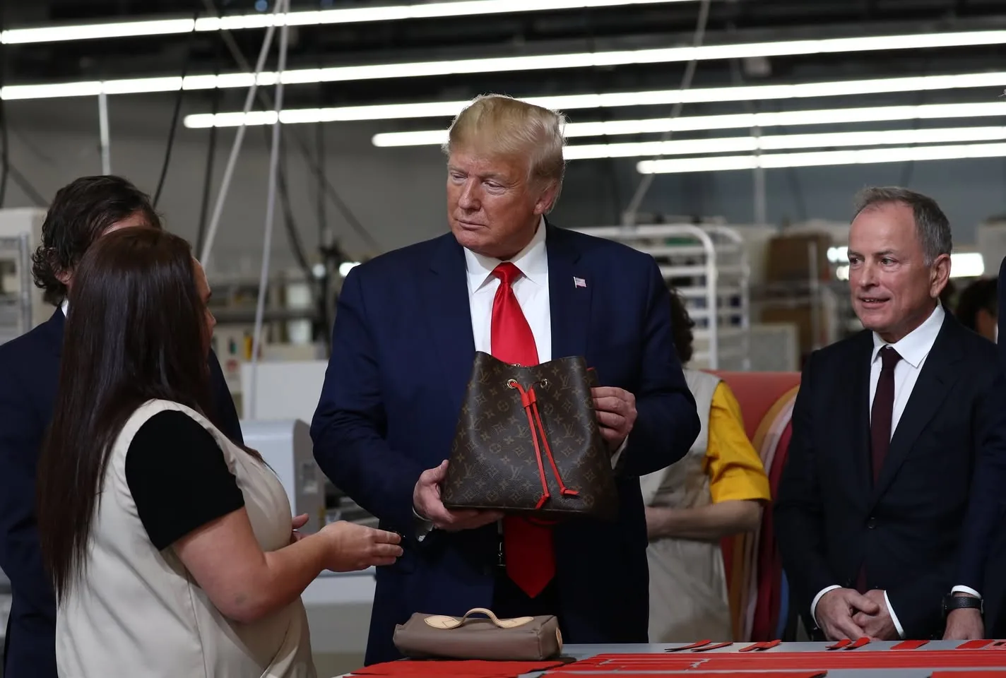 The image depicts a group of individuals in a factory or workshop setting, characterized by bright overhead lighting and industrial equipment in the background. 

In the center, a man, possibly a public figure, is holding a handbag. The bag is brown with a distinctive pattern that resembles the Louis Vuitton logo, featuring a red drawstring. He is wearing a dark blue suit with a white shirt and a bright red tie, and he has light-colored hair. His expression appears focused as he looks at the handbag.

To his left, a woman is engaged in conversation with him. She has long brown hair and is wearing a black shirt with a light-colored vest. She is gesturing with her hands, indicating she is explaining something about the bag. 

To the right of the man, another individual in a dark suit is smiling and looking at the man holding the bag. This person has short hair and is wearing a white shirt with a dark tie. 

In the background, there are several people in light-colored uniforms, suggesting they are workers in the facility. The setting appears to be a production area, with machinery and materials visible, indicating an operational environment related to manufacturing or assembly.

The table in front of them has various items, including another smaller bag that is beige in color, and several red items, possibly fabric or components related to the production process. The overall atmosphere seems to be one of a business or promotional event, focused on showcasing products.