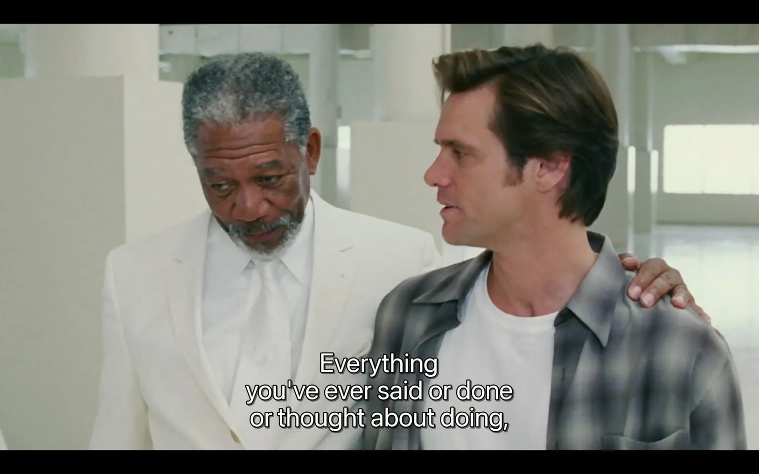 The image features two men engaged in conversation. 

**Readable Text:**
- The text displayed at the bottom of the image reads: "Everything you've ever said or done or thought about doing," which appears to be part of a dialogue.

**People:**
1. **First Man:**
   - He is wearing a white suit, which includes a white jacket and a white tie. 
   - His shirt is also white, and he has a light gray beard and hair that is slightly curly and graying.
   - His facial expression appears thoughtful, with his gaze directed slightly downward.
   - He has a calm demeanor and is standing close to the second man.

2. **Second Man:**
   - He is dressed in a casual outfit consisting of a white t-shirt under an unbuttoned gray and black plaid shirt.
   - His hair is dark brown and styled in a slightly tousled manner.
   - He has a slight smile and is looking at the first man, suggesting engagement in the conversation.
   - He has his right hand resting on the shoulder of the first man.

**Setting:**
- The background is a bright, spacious interior, likely a modern building or studio. 
- The walls are white, and there are large windows or openings that allow natural light to flood the space.
- The floor appears to be smooth and light-colored, contributing to the overall bright ambiance.
- There are no visible objects or furniture in the immediate background, emphasizing the focus on the two men.

**Environmental Context:**
- The setting seems to be a professional or artistic environment, possibly indicating a scene from a film or a creative meeting space.
- The overall atmosphere is open and airy, with a minimalist design that draws attention to the subjects in the foreground.

This description captures the observable details without interpretation or speculation.