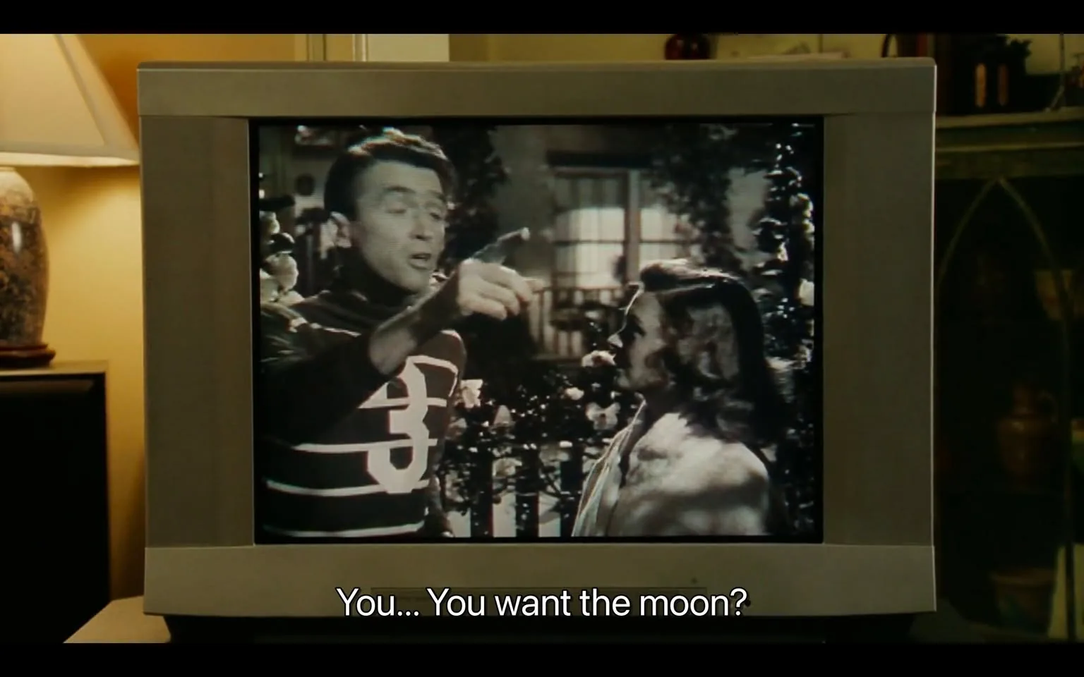 The image depicts a scene from a black-and-white film displayed on a vintage television set. The screen shows two characters engaged in conversation. 

**Television and Screen:**
- The television is a boxy, older model with a thick frame, likely from the mid-20th century. The screen displays a scene from a film, which is in black and white.
- The visible text at the bottom of the screen reads: "You... You want the moon?" indicating a dialogue from the film.

**Characters:**
- The character on the left is a man with short, slicked-back hair. He is wearing a dark sweater with horizontal white stripes and a large number "3" prominently displayed on the front. His expression appears animated as he gestures with his right hand, pointing while speaking.
- The character on the right is a woman with medium-length, wavy hair. She is wearing a light-colored garment that appears to be a shawl or wrap. Her facial expression is attentive, and she is looking at the man as he speaks.

**Background:**
- The background of the scene includes a garden-like setting, with what appears to be a fence and some foliage, suggesting an outdoor scene. There are hints of a house or building visible through the background, with windows that are partially obscured.
- The lighting in the scene is soft, typical of classic films, enhancing the vintage aesthetic.

**Surrounding Environment:**
- To the left of the television, there is a lamp with a beige lampshade, providing ambient light to the room. The base of the lamp appears to be ceramic or glass, with a decorative pattern.
- In the background, there are shelves or furniture pieces that hold various items, possibly decorative objects or plants, but specific details are not discernible.

Overall, the image captures a moment from a classic film, emphasizing the characters' interaction and the nostalgic feel of the setting.