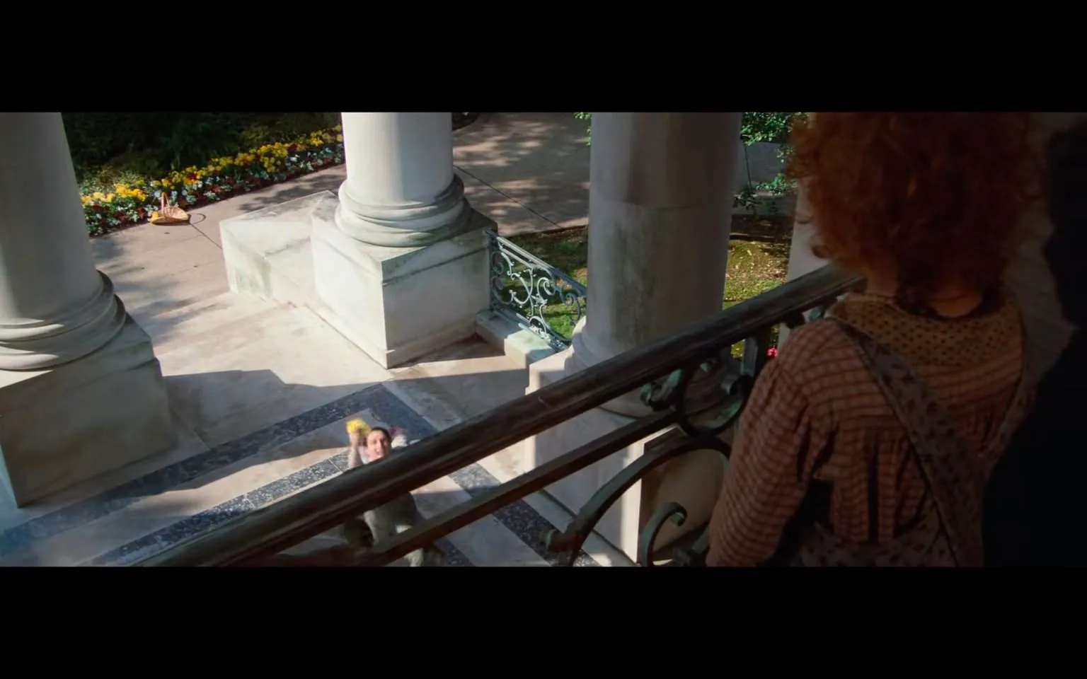 The image depicts a scene from a film or a staged setting, featuring a balcony view overlooking a garden area. 

In the foreground, there is a young girl with curly red hair, wearing a brown dress with a checkered pattern and a light-colored apron. She is positioned on the balcony, facing downwards. The girl appears to be observing or interacting with someone below, but her facial expression is not clearly visible due to the angle.

Below the balcony, two individuals are visible on the steps. One person is dressed in a light-colored outfit, possibly a dress or a costume, and is holding a yellow object, which may be a toy or a prop. This person is looking up towards the balcony, suggesting a moment of engagement or playfulness. The second individual is partially obscured but appears to be wearing a darker outfit.

The background features a well-maintained garden with a variety of colorful flowers arranged in neat rows along a pathway. The flowers are predominantly yellow, with hints of other colors, contributing to a vibrant outdoor setting. The ground is paved, and there are stone steps leading up to the balcony area.

The architectural elements include large, white columns that support the balcony, with intricate ironwork visible in the railing. The overall setting suggests a historical or grand location, possibly a mansion or a public building, given the classical architectural style.

No readable text, signs, or logos are present in the image. The focus remains on the characters and the surrounding environment, with no identifiable brands or trademarks visible. The lighting appears natural, indicating that the scene is set during the day.
