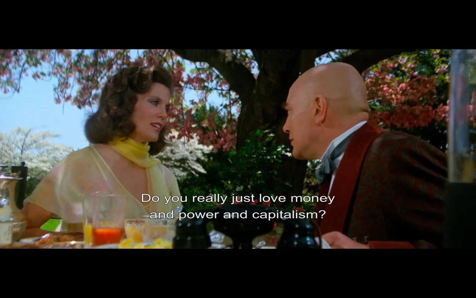 The image depicts a scene from a film featuring two characters engaged in conversation. 

**Readable Text:**
- The text visible at the bottom of the image reads: "Do you really just love money and power and capitalism?"

**Characters:**
1. **Female Character:**
   - She has medium-length, wavy brown hair styled with volume.
   - Her outfit consists of a light, sheer yellow dress with a deep neckline and a matching yellow scarf around her neck.
   - She is positioned slightly to the left, facing the male character, and appears to be in mid-conversation, with a serious expression on her face.

2. **Male Character:**
   - He is bald and has a fair complexion.
   - He wears a formal outfit that includes a patterned brown jacket with a high collar and a light blue shirt underneath.
   - His facial expression is focused as he listens to the female character, and he is slightly turned towards her, indicating engagement in the conversation.

**Setting:**
- The background features a vibrant outdoor scene with a tree adorned with pink blossoms, suggesting a spring or summer setting.
- The table in front of the characters is set for a meal, with various items visible:
  - A glass pitcher, possibly for juice or water.
  - Several glasses, one containing a reddish liquid, and another with a yellow drink.
  - A plate with food items, though specifics are not discernible.
  - Two black pepper shakers are prominently displayed on the table.

**Environmental Context:**
- The scene appears to take place in a garden or outdoor dining area, indicated by the greenery and flowering tree.
- The overall atmosphere is bright and colorful, contributing to a lively setting.

The image captures a moment of dialogue between the two characters, with a focus on their attire and the surrounding natural beauty.