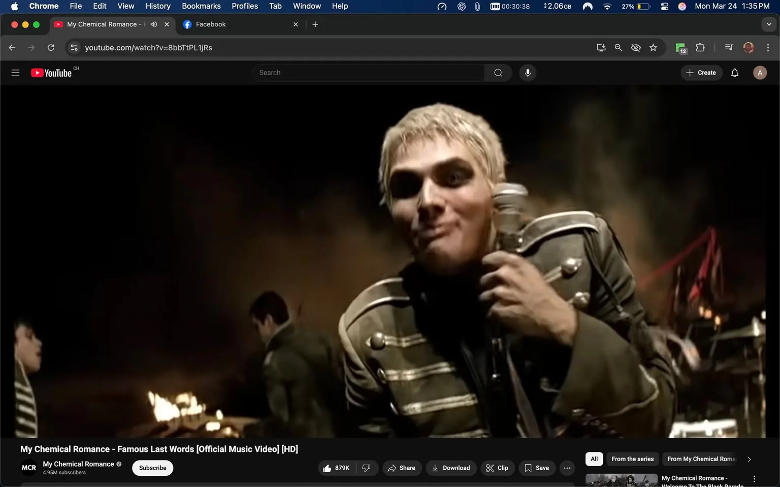 The image depicts a scene from the music video "Famous Last Words" by My Chemical Romance. In the foreground, a male figure with short, platinum blonde hair is prominently featured. He is holding a microphone in his right hand and appears to be singing or performing. His facial expression is somewhat playful or mischievous, with a slight smirk. He is wearing a military-style jacket that is dark green or gray with silver stripes and buttons, giving it a distinctive, structured look. The jacket has a high collar and appears to be fitted.

In the background, another male figure is partially visible, dressed similarly in a military-style jacket. This figure is positioned to the left of the main subject and is facing away from the camera, suggesting he is engaged in an activity related to the performance. The setting is dark and smoky, with a blurred background that includes flames or fire, indicating a dramatic or intense atmosphere. The lighting is low, contributing to a moody ambiance.

The video interface at the bottom of the image shows the title "My Chemical Romance - Famous Last Words [Official Music Video] [HD]" along with the channel name "My Chemical Romance" and a subscriber count of 4.95 million. There are also options for subscribing, sharing, downloading, and clipping the video, which are typical features of a YouTube interface.

Overall, the image captures a moment from a high-energy performance, characterized by the band's signature aesthetic, which often includes themes of rebellion and emotional expression. The visual elements, including the clothing and setting, align with the band's style and the tone of the music video.