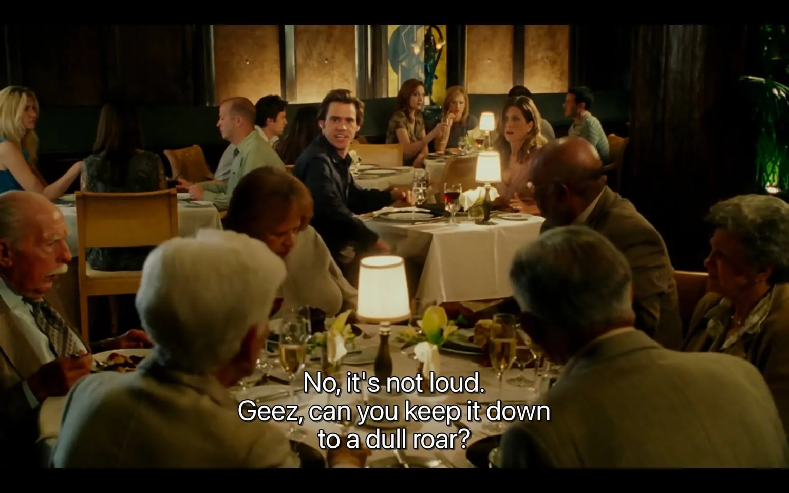 The image depicts a restaurant scene filled with diners seated at tables. The setting is well-lit, with soft lighting provided by small lamps on each table. The tables are covered with white tablecloths, and each table is set with plates, glasses, and utensils, indicating a formal dining experience.

In the foreground, a group of older adults is seated at a table. One man, with a bald head and a prominent mustache, is wearing a light-colored suit and appears to be engaged in conversation while holding a fork. Next to him, a woman with short, gray hair is dressed in a light-colored blouse and is looking down at her plate. Another older man, wearing glasses and a dark suit, is seated across from them, seemingly listening intently.

In the background, a younger man with dark hair and a blue shirt is looking towards the camera, displaying a surprised or concerned expression. He is seated at a table with a woman who has long, dark hair and is wearing a black dress. Another woman, dressed in a blue dress, is seated nearby, engaged in conversation with a man in a green shirt.

The restaurant has a modern decor, with dark wood paneling and abstract art visible on the walls. The atmosphere appears lively, with multiple groups of diners engaged in conversation. The text overlay at the bottom of the image reads: "No, it's not loud. Geez, can you keep it down to a dull roar?" This suggests a humorous or light-hearted exchange occurring in the scene.

Overall, the image captures a bustling dining environment, showcasing a mix of ages and styles among the patrons, with a focus on the interactions and expressions of the individuals present.