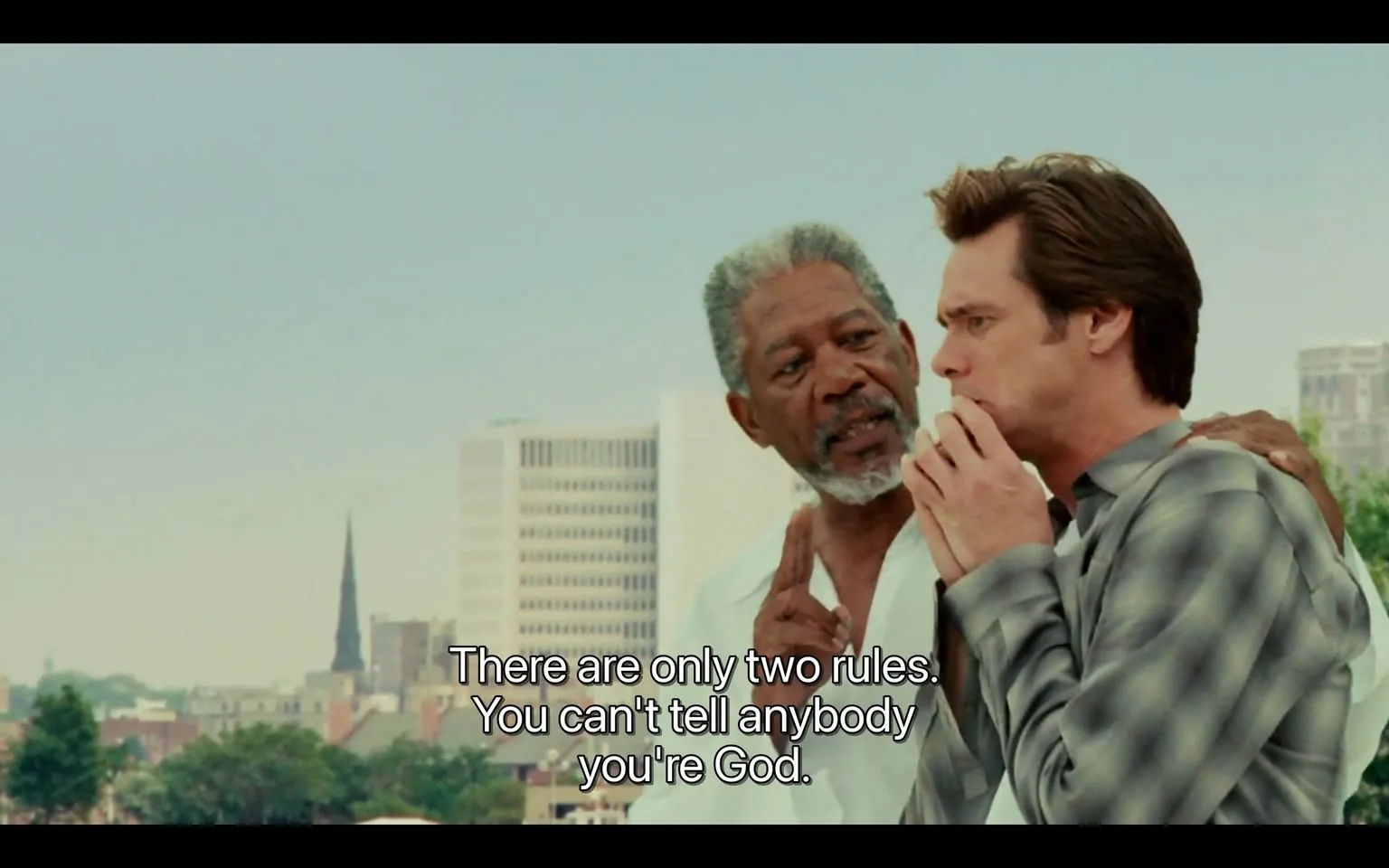 The image depicts two men engaged in conversation against a cityscape backdrop. 

**Readable Text:**
- There is a caption at the bottom of the image that reads: "There are only two rules. You can't tell anybody you're God."

**People:**
1. **First Man:**
   - He has dark skin and gray hair, with a beard and mustache. 
   - He is wearing a white shirt, which appears to be unbuttoned at the collar, giving a relaxed appearance.
   - His facial expression seems serious, and he is looking intently at the second man.
   - He has a hand on the second man's shoulder, suggesting a guiding or mentoring gesture.

2. **Second Man:**
   - He has light skin and brown hair, styled in a slightly tousled manner.
   - He is wearing a gray plaid shirt, which is buttoned up.
   - His facial expression appears contemplative or concerned, and he is holding his chin with one hand, indicating deep thought.
   - He is looking towards the first man, suggesting engagement in the conversation.

**Setting:**
- The background features a city skyline with several buildings, including a tall structure that has a spire, possibly a church or a tower.
- The sky is clear with a light blue hue, indicating a sunny day.
- There are green trees visible in the foreground, adding a natural element to the urban setting.

**Environmental Context:**
- The scene appears to be set outdoors, likely on a rooftop or elevated area, given the perspective of the city below.
- The overall atmosphere seems to be casual yet serious, as indicated by the body language and expressions of the two men.

This description captures the observable details in the image without speculation or subjective interpretation.