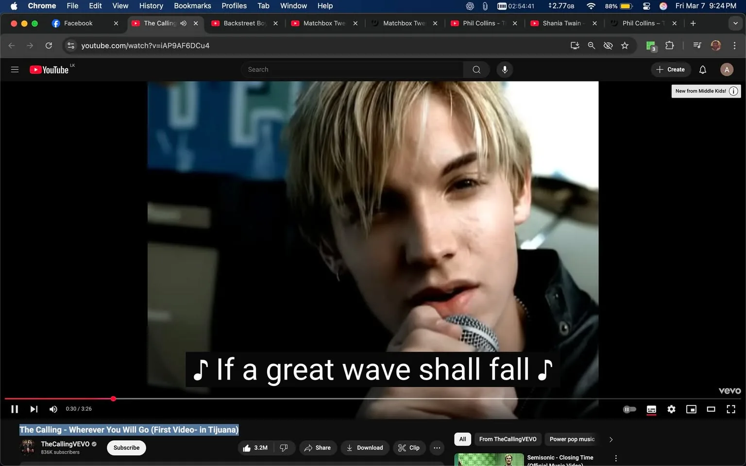 The image shows a scene from a music video on YouTube, specifically from the song "Wherever You Will Go" by The Calling. The video is currently playing, and the timestamp indicates it is at 0:30 out of a total of 3:26 minutes. 

In the foreground, a young male singer is prominently featured. He has light blonde hair styled in a slightly tousled manner, with strands falling across his forehead. His facial expression appears focused and intense as he holds a microphone close to his mouth. He is wearing a black jacket, which is partially unzipped, revealing a light-colored shirt underneath. The background is somewhat blurred but suggests a performance setting, possibly with a stage or band equipment visible.

The video includes subtitles at the bottom, displaying the lyrics: "If a great wave shall fall." The text is in a white font against a black background, making it easily readable. 

The YouTube interface is visible at the top of the image, showing the video title, "The Calling - Wherever You Will Go (First Video- in Tijuana)," along with the channel name "TheCallingVEVO" and its subscriber count of 836K. Below the video, there are options to subscribe, share, and download, along with a list of related videos.

The overall setting appears to be a music video shoot, likely in an urban environment, given the hints of band equipment and the casual attire of the performer. The lighting is bright, focusing on the singer, while the background remains less illuminated, emphasizing his presence.