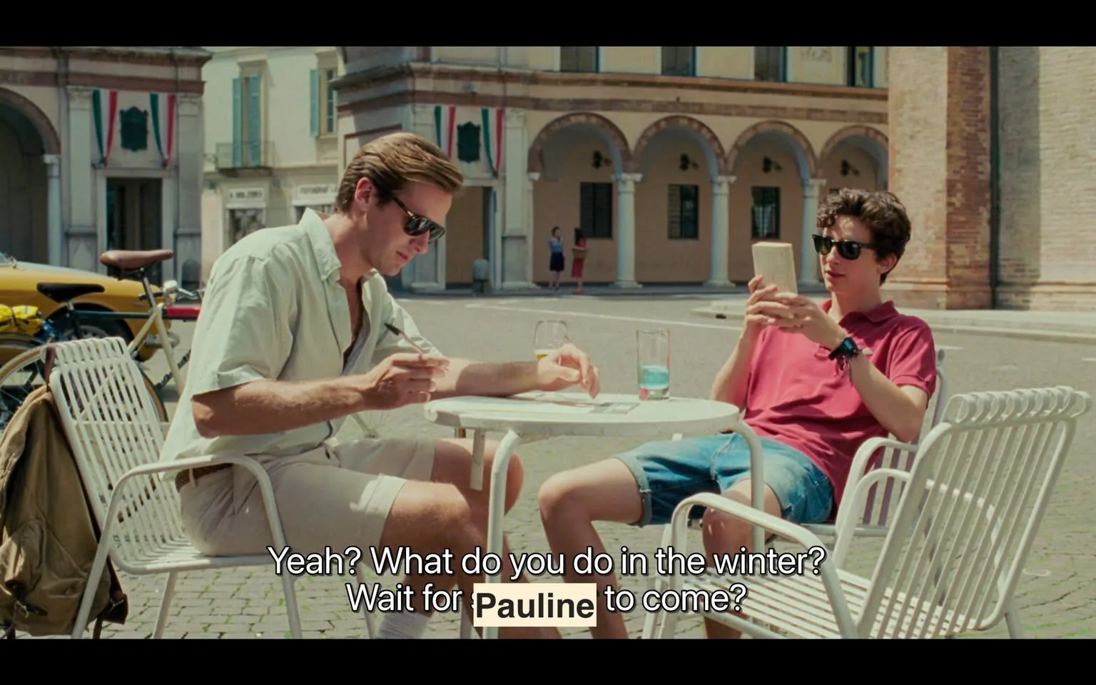 The image depicts two young men seated at a small round table in an outdoor café setting. The table is white and has two glasses on it—one appears to contain a clear liquid, possibly water, while the other is empty. 

The man on the left has light brown hair styled back and is wearing dark sunglasses. He is dressed in a light-colored, short-sleeved button-up shirt that is unbuttoned at the collar, and white shorts. He is holding a pen and appears to be writing or drawing on a piece of paper on the table. His expression is focused as he looks down at the paper.

The man on the right has curly dark hair and is also wearing dark sunglasses. He is dressed in a short-sleeved, red polo shirt and blue denim shorts. He is holding a smartphone in both hands and is looking at it, seemingly engaged with whatever is on the screen. 

In the background, there are several buildings with arched doorways and columns, suggesting a historical or picturesque setting. The architecture features light-colored walls and some decorative elements. There are also Italian flags visible, indicating the location may be in Italy. 

To the left of the table, a bicycle is parked, and there is a yellow vehicle visible in the background. The ground is paved with cobblestones, and the overall atmosphere appears relaxed and casual, typical of a sunny day in a European town. 

No other people are prominently visible in the immediate vicinity, although a few figures can be seen in the background, contributing to the lively ambiance of the scene.