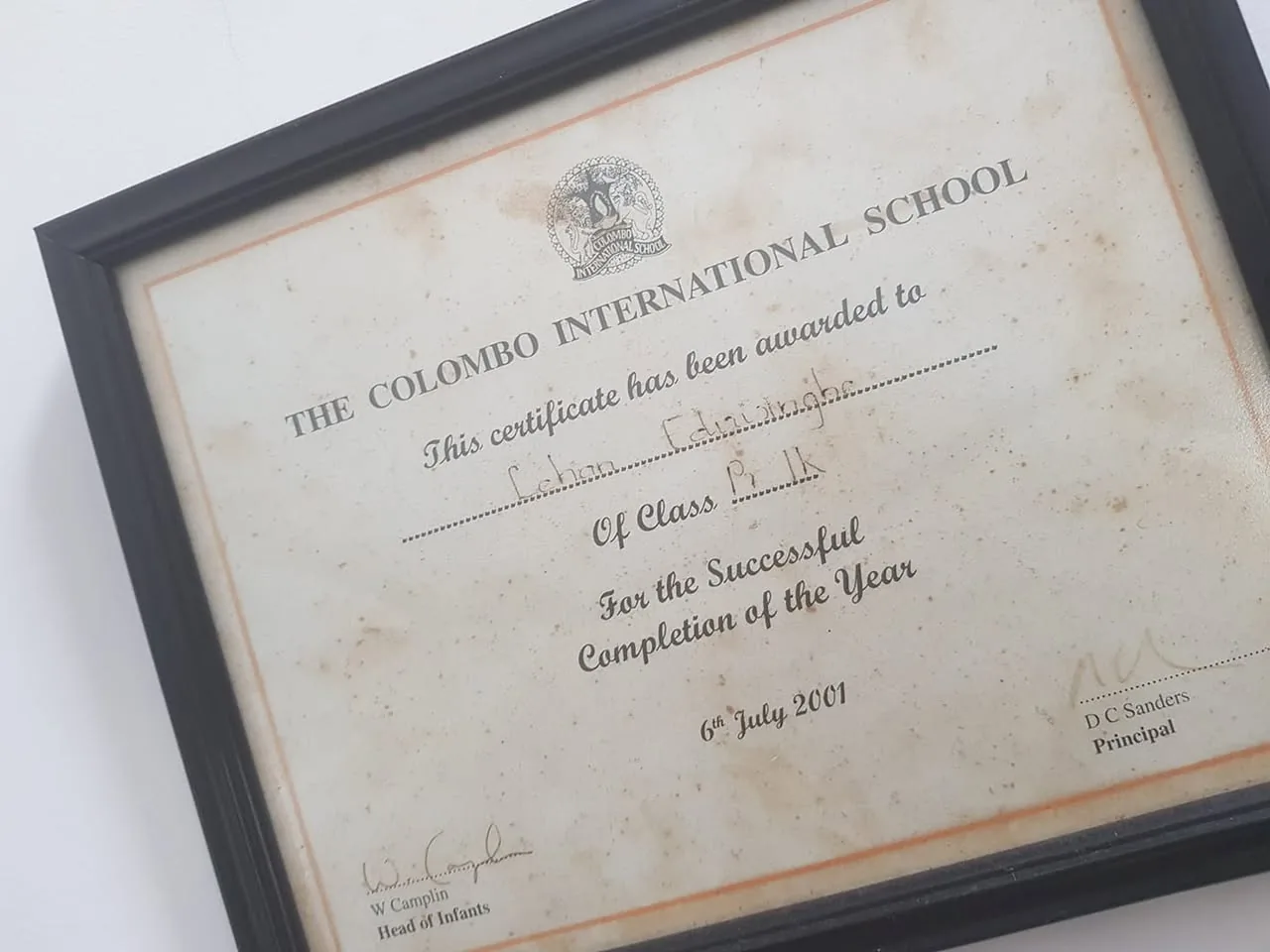 The image displays a certificate framed in a black frame. The certificate is titled "THE COLOMBO INTERNATIONAL SCHOOL" and is printed on a slightly yellowed, textured paper, indicating some age. 

At the top, there is a logo or emblem, likely representing the school, but the details are not clearly visible. Below the title, the text reads, "This certificate has been awarded to," followed by a handwritten name that appears to be "Lehan Edirisinghe." The name is written in a cursive style, suggesting a personal touch.

The certificate specifies "Of Class P.1 K," indicating the class level of the recipient. Below this, it states, "For the Successful Completion of the Year," which signifies the purpose of the award. The date at the bottom reads "6th July 2001," marking when the certificate was issued.

The signatures of two individuals are present at the bottom: one appears to be "W. Camplin," noted as "Head of Infants," and the other is "D C Sanders," identified as "Principal." The signatures are handwritten, adding to the authenticity of the document.

The background of the certificate is plain, with a white wall visible behind the frame, and there are some smudges and stains on the certificate itself, suggesting it has been handled over time. The frame is simple and rectangular, with no additional decorations or embellishments. 

Overall, the certificate serves as a formal acknowledgment of achievement from the Colombo International School, capturing a moment in the recipient's educational journey.