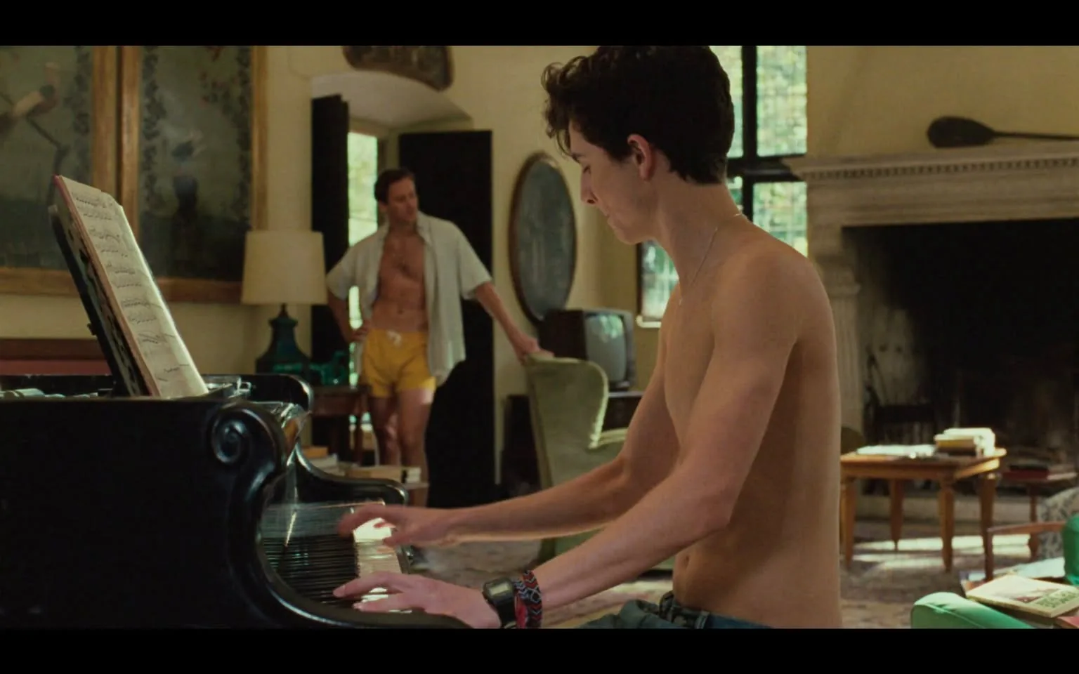 The image depicts an interior scene featuring two individuals in a room with a warm, inviting atmosphere. 

In the foreground, a young man is seated at a black grand piano, playing music. He is shirtless, with short, curly hair and a slender build. His left hand is positioned over the piano keys, while his right hand is slightly raised, possibly preparing to play. He is wearing denim shorts and a colorful wristband on his left wrist. His expression is focused, suggesting concentration on the music.

In the background, another man stands near a doorway. He has a muscular build and is wearing yellow shorts, with a white, unbuttoned shirt that hangs loosely over his torso. His hair is dark and slightly tousled. He appears to be looking at the young man at the piano, with a relaxed posture, hands on his hips.

The room features large windows that allow natural light to filter in, illuminating the space. There are decorative elements on the walls, including framed artwork that depicts figures, and a large mirror. A lamp stands on a side table next to the piano, and there is a green armchair visible in the background. A television and some books are also present on a nearby table, contributing to the cozy ambiance of the setting. The floor is covered with a patterned rug, adding warmth to the room.

Overall, the scene conveys a sense of intimacy and creativity, with the piano serving as a focal point in the space.