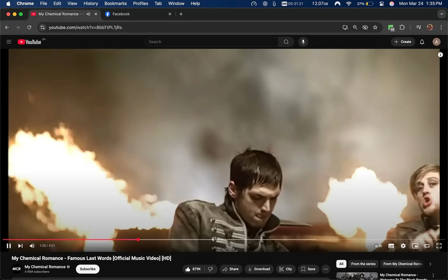 The image displays a scene from a music video by My Chemical Romance, specifically titled "Famous Last Words." The video is playing on a YouTube interface, which is visible at the top of the image. The YouTube logo is present, along with the video title and channel name, "My Chemical Romance," which has 4.95 million subscribers. Below the video, there are options to like, share, download, and clip the video, with a visible like count of 879,000.

In the foreground, two male figures are prominently featured. The figure on the left has short, dark hair and is wearing a military-style jacket with a high collar and multiple buttons. His expression appears serious and focused as he looks downward. The second figure, positioned to the right, has slightly longer, light-colored hair and is wearing a similar military-style jacket. He is facing the camera and appears to be singing or expressing emotion, with his mouth open.

The background is filled with flames, creating a dramatic and intense atmosphere. The flames are bright and orange, contrasting against a blurred, smoky backdrop that suggests an explosive or chaotic setting. The overall lighting is dim, with the flames providing the primary source of illumination.

The video is currently at 1 minute and 20 seconds into its 4-minute and 21-second runtime. The interface shows the video controls, including a play/pause button, volume control, and a timeline slider. The browser window is open, indicating that the image was captured on a computer, with additional tabs visible for Facebook and other browsing activities. 

No other text, logos, or identifiable brands are present in the image aside from those related to YouTube and My Chemical Romance. The overall composition conveys a sense of urgency and intensity, typical of the band's style and thematic elements in their music videos.