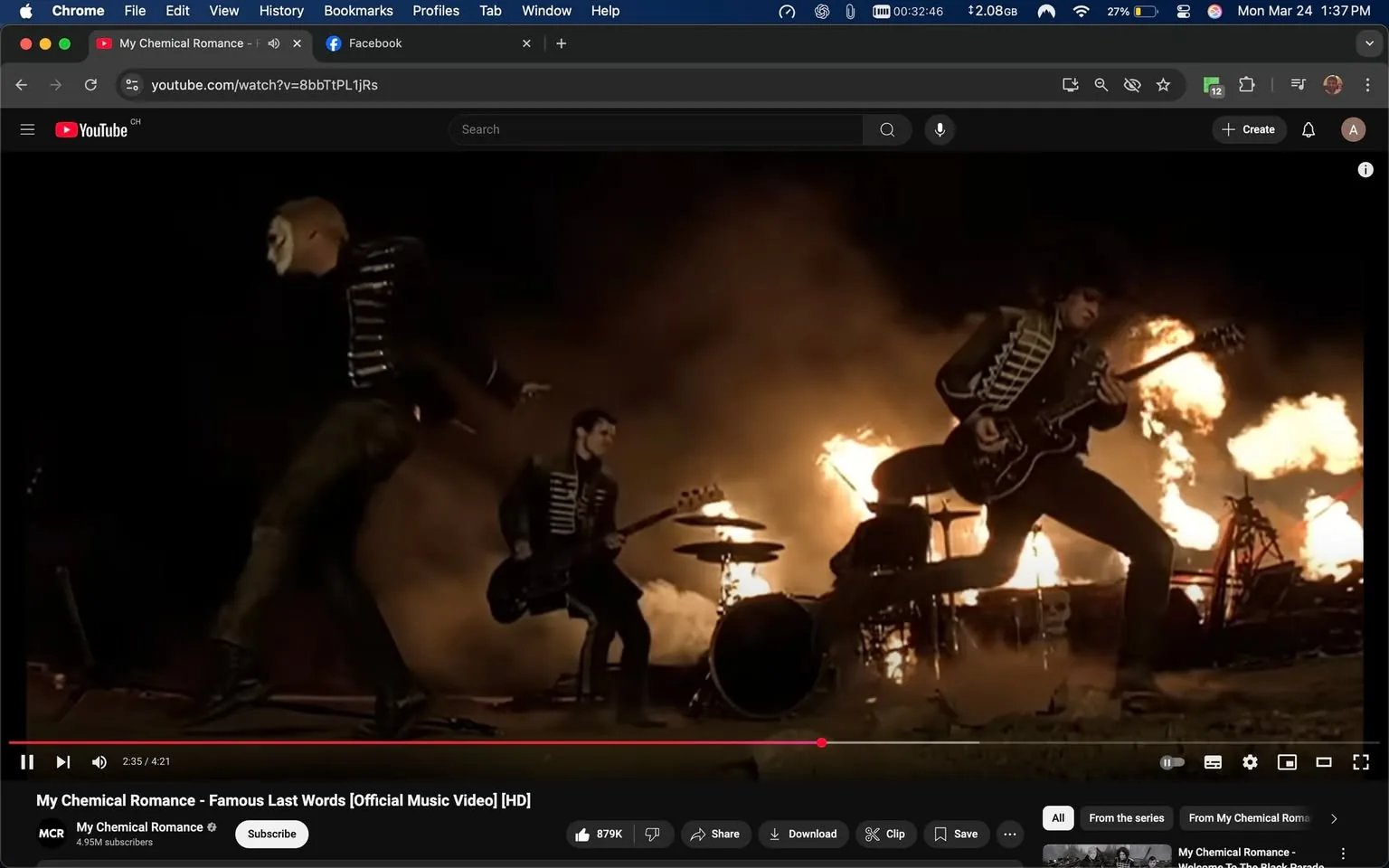 The image depicts a scene from the music video for "Famous Last Words" by My Chemical Romance. The setting appears to be an outdoor performance, likely at night, with a dramatic backdrop of flames and smoke, creating an intense atmosphere.

In the foreground, three band members are visible. The first member on the left is wearing a black military-style jacket with white stripes and has a white face paint design, resembling a skull. They are in a dynamic pose, possibly mid-performance, with their body angled and one arm raised, pointing outward.

The second member, positioned centrally, is playing a black bass guitar. They are dressed similarly in a black jacket with white stripes and have a serious expression on their face. The drummer is partially obscured behind the drum set, which consists of a black bass drum and other percussion instruments. The drummer's silhouette is visible against the flames, but their features are not clearly discernible.

The third member, on the right, is in a mid-jump stance, playing an electric guitar. They are also wearing a black jacket with white stripes. The flames in the background are bright orange and yellow, contrasting sharply with the dark clothing of the band members. The ground appears to be dirt or gravel, and the overall lighting is low, enhancing the dramatic effect of the fire.

In the bottom portion of the image, there is a YouTube interface visible, showing the video title "My Chemical Romance - Famous Last Words [Official Music Video] [HD]" along with the channel name "My Chemical Romance" and a subscriber count of 4.95M. The video progress bar indicates a timestamp of 2:35 out of a total duration of 4:21. There are also buttons for subscribing, sharing, downloading, and other video options.

Overall, the image captures a high-energy moment from a rock performance, characterized by the band's theatrical style and the dramatic visual effects of fire and smoke.
