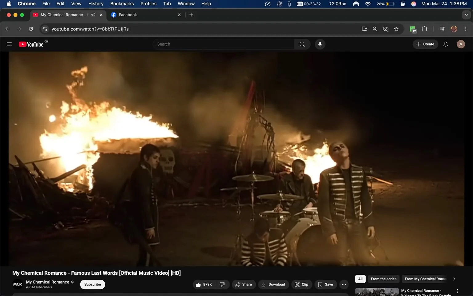 The image depicts a scene from the music video "Famous Last Words" by My Chemical Romance. In the foreground, there are four male figures, all dressed in dark, military-style clothing. The first figure on the left is wearing a black jacket with white stripes and appears to be looking down. He has a dark hairstyle and is holding a bag slung over his shoulder. The second figure, who is seated on the ground, is wearing a black and white striped shirt and has a distressed expression, with his hands clasped together in front of him. The third figure, positioned behind the drums, is wearing a black jacket and has a serious expression. The fourth figure, slightly to the right, is standing with his head tilted back and eyes closed, wearing a black jacket with metallic embellishments.

In the background, there is a large fire burning, creating a dramatic and intense atmosphere. The flames illuminate the scene, casting a warm glow on the figures. Behind the fire, there is a structure that appears to be partially collapsed, with a large skull painted on it, adding to the overall dark aesthetic of the setting. The ground is uneven and appears to be dirt or gravel, contributing to the rugged environment.

The video interface at the bottom of the image includes the YouTube logo and the title "My Chemical Romance - Famous Last Words [Official Music Video] [HD]." There are also buttons for subscribing, saving, and downloading, along with a view count of 879K. The branding for My Chemical Romance is visible, indicating the band's presence and popularity. The overall mood of the scene is somber and intense, consistent with the themes often explored in the band's music.