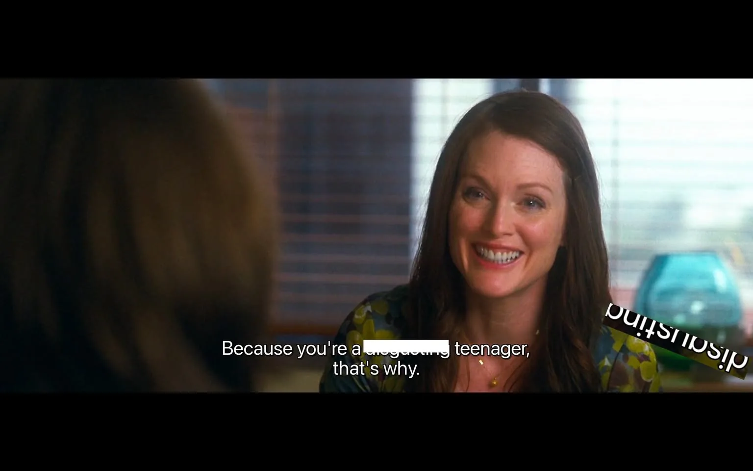 The image depicts a scene from a film featuring a woman with long brown hair, styled in loose waves. She is smiling broadly, showcasing a friendly and engaging demeanor. The woman is wearing a colorful, patterned blouse with shades of green, yellow, and blue, which adds a vibrant touch to her appearance. Her facial expression conveys warmth and approachability, as she appears to be in a conversation.

In the background, there are horizontal blinds partially obscuring a window, allowing soft light to filter into the room. The setting appears to be an indoor space, likely an office or a meeting room, given the presence of a desk and the formal nature of the conversation. There is a turquoise glass object on the table, possibly a decorative piece, which adds a pop of color to the environment.

The visible text on the screen reads: "Because you're a [censored] teenager, that's why." This dialogue suggests a playful or teasing tone in the conversation, although the context is not fully clear from the image alone. The text is displayed in white font against a dark background, making it easily readable.

Overall, the scene captures a moment of interaction between characters, with a focus on the woman's expressive face and the casual yet colorful setting.