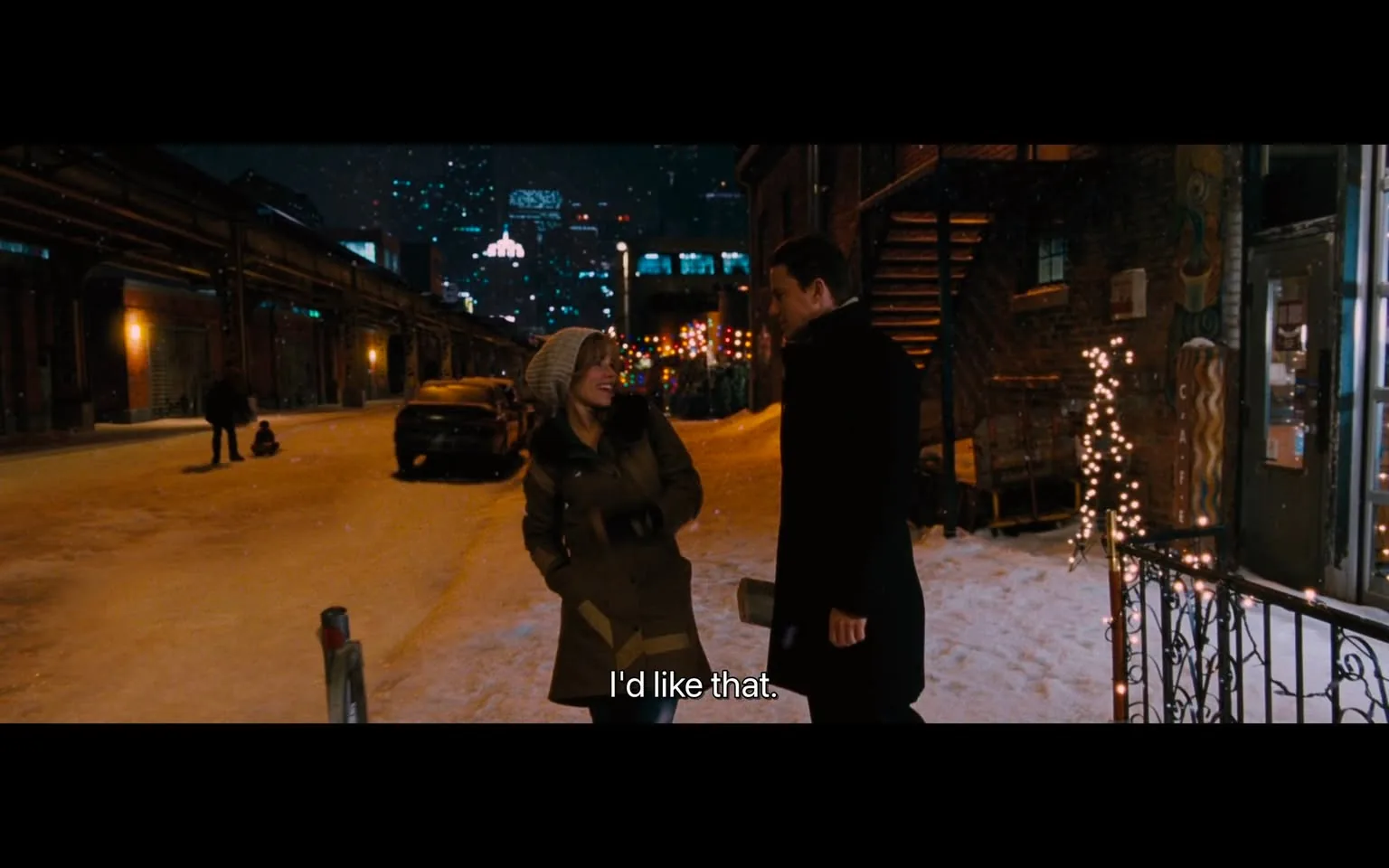 The image depicts a nighttime urban scene with a snowy street. In the foreground, two individuals are engaged in conversation. The first person, a woman, is wearing a light-colored coat with a fur-lined hood and a knitted beanie. She has a cheerful expression, smiling as she looks at the second person. The second person, a man, is dressed in a dark coat and appears to be smiling back at her. They are standing close together, suggesting a friendly or intimate interaction.

In the background, the street is lined with buildings that have a mix of brick and modern architecture. There are decorative lights strung along the buildings, creating a festive atmosphere. Snow covers the ground, and a few parked cars are visible, one of which is partially covered in snow. In the distance, there are more buildings with illuminated windows, contributing to the urban night scene. 

The overall ambiance is cozy and inviting, enhanced by the snowfall and the warm glow of the lights. The setting appears to be a quiet street, possibly in a city known for its winter charm. No specific signs, logos, or brands are visible in the image. The focus remains on the two individuals and their interaction against the backdrop of the snowy urban environment.