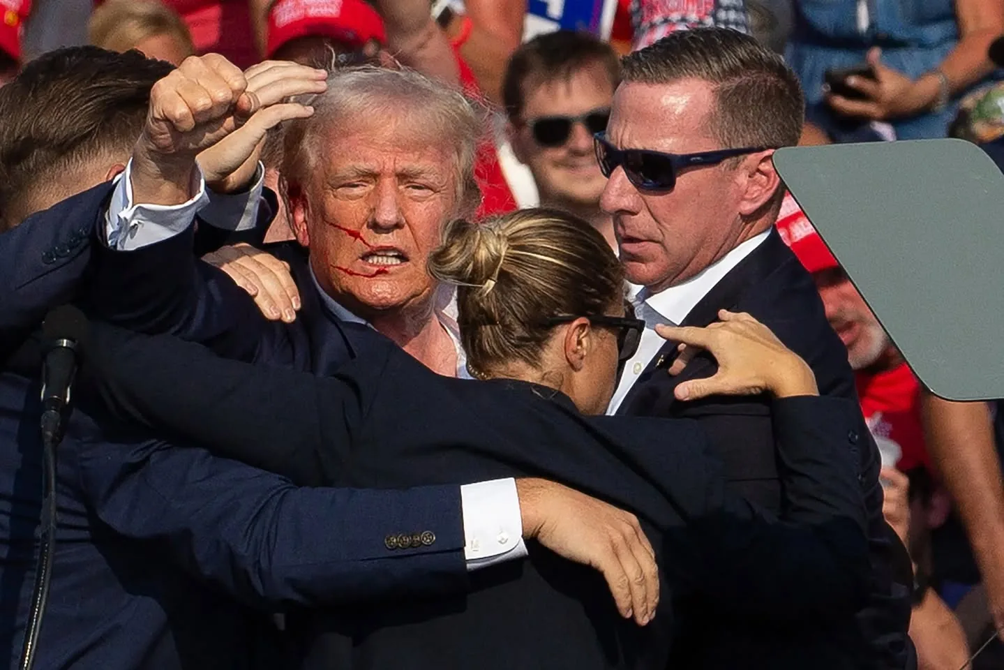 The image depicts a chaotic scene involving a group of individuals surrounding a central figure, who appears to be a man with distinct features. He has light-colored hair and is wearing a dark suit with a white shirt. His face shows visible signs of distress, including a noticeable red mark or streak across his face, suggesting he may have been injured. The expressions on his face convey a sense of urgency or alarm.

Surrounding him are several individuals, some of whom are dressed in dark suits, indicating a formal setting, possibly a political rally or event. Their body language suggests they are providing protection or assistance to the central figure. One person, a woman with her hair tied back, is positioned close to him, while others are reaching out, possibly trying to manage the situation.

In the background, a crowd is visible, with many individuals wearing red hats, which may indicate support for a specific political affiliation. The setting appears to be outdoors, with a stage or podium visible, suggesting this is a public event. The crowd's expressions and posture indicate they are engaged and possibly reacting to the unfolding situation.

No readable text, signs, or logos are clearly visible in the image. The overall atmosphere conveys a moment of heightened tension, with the central figure at the focus of the surrounding individuals.