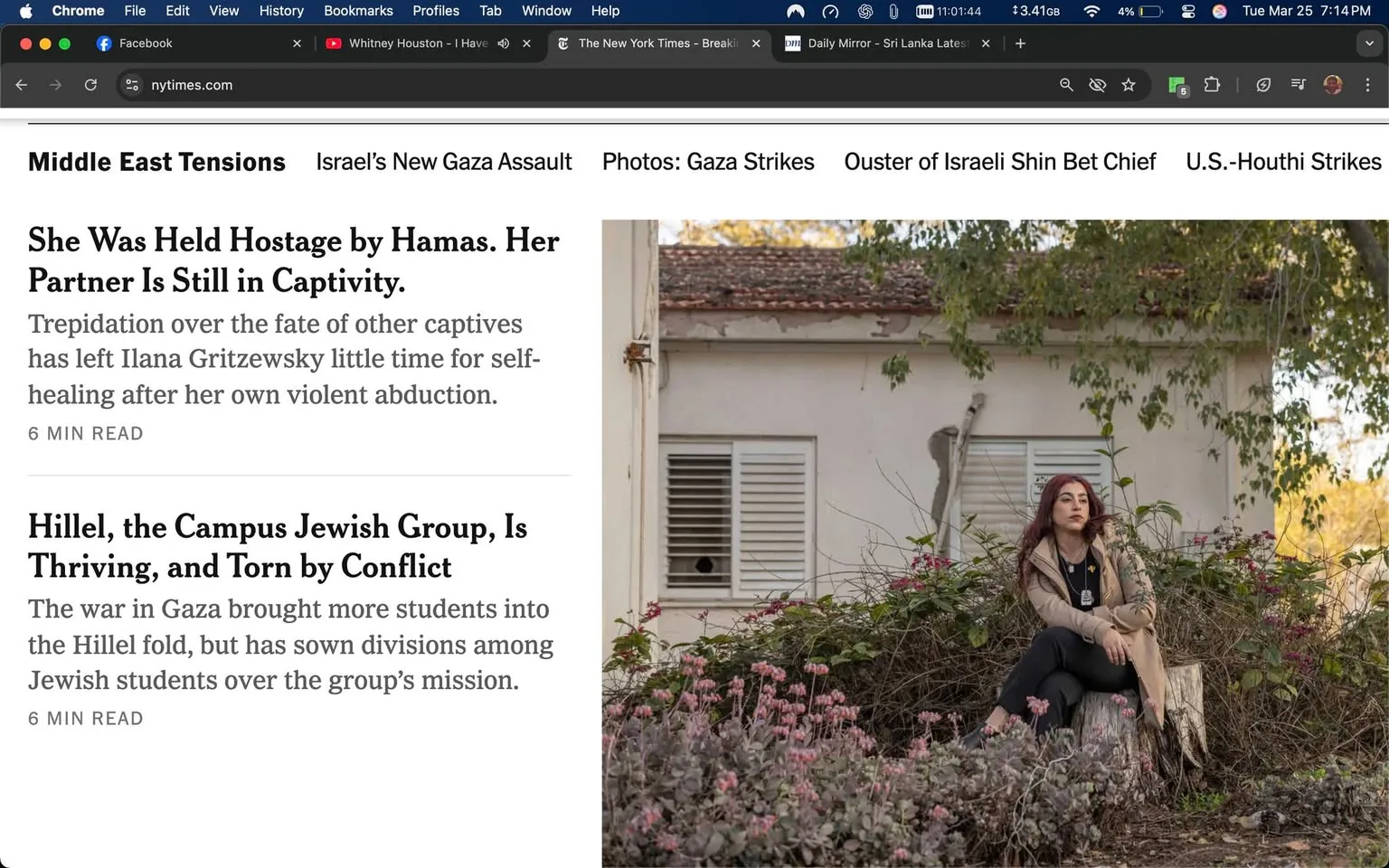 The image is a screenshot of a webpage from The New York Times, specifically focusing on articles related to Middle East tensions. 

**Text Content:**
- The header reads "Middle East Tensions" followed by "Israel's New Gaza Assault."
- The main article title is "She Was Held Hostage by Hamas. Her Partner Is Still in Captivity." Below this, there is a brief description: "Trepidation over the fate of other captives has left Ilana Gritzewsky little time for self-healing after her own violent abduction." This section indicates a reading time of "6 MIN READ."
- Another article titled "Hillel, the Campus Jewish Group, Is Thriving, and Torn by Conflict" is also visible, with a description stating: "The war in Gaza brought more students into the Hillel fold, but has sown divisions among Jewish students over the group’s mission." This section also indicates a reading time of "6 MIN READ."

**Visual Elements:**
- The right side of the image features a photograph of a woman sitting on a log in a garden-like setting. 
- She has long, wavy hair that appears to be reddish-brown. 
- Her clothing consists of a light-colored, possibly beige, jacket over a black top, paired with black pants. 
- She is seated with her arms crossed, looking directly at the camera with a neutral expression.
- The background includes a white building with a sloped roof, partially obscured by greenery. There are several plants and flowers in the foreground, adding color to the scene.

**Setting:**
- The environment appears to be outdoors, possibly in a garden or park, with natural light illuminating the scene.
- The building in the background has windows with shutters, and there are visible signs of wear, such as peeling paint.

Overall, the image captures a blend of textual information about current events and a poignant visual representation of an individual in a serene yet potentially reflective setting.