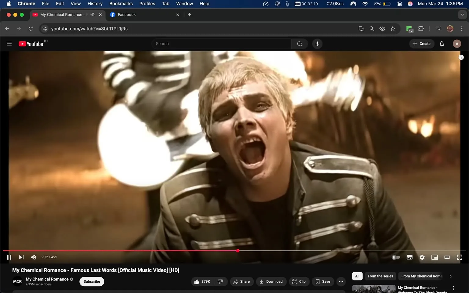 The image is a still from a music video by My Chemical Romance, specifically from the song "Famous Last Words." 

**Readable Text:**
- The video player interface at the bottom shows the title "My Chemical Romance - Famous Last Words [Official Music Video] [HD]" and the duration of the video (2:12 out of 4:21). 
- The YouTube logo is visible in the top left corner, along with the channel name "My Chemical Romance" and the subscriber count of 4.95M.

**Visible Brands and Objects:**
- The video is hosted on YouTube, indicated by the logo and interface elements.
- There are electric guitars in the background, with one prominently featured, appearing white.

**People's Clothing and Appearance:**
- The central figure is a male performer with short, spiky blonde hair. 
- He is wearing a dark military-style jacket with silver embellishments and buttons. The jacket has a high collar and is fitted, giving a structured appearance.
- His facial expression is intense, with his mouth open in a scream or shout, showcasing a strong emotional expression.
- The performer has dark eye makeup, enhancing the dramatic look typical of the band's aesthetic.

**Setting and Background:**
- The background is dimly lit, with warm tones suggesting a performance setting, possibly a stage or a rehearsal space.
- There are blurred figures in the background, likely other band members, dressed in similar military-style attire, contributing to a cohesive visual theme.
- The environment appears to be energetic and dynamic, typical of a rock music performance.

Overall, the image captures a moment of high emotion and intensity, characteristic of the band's performance style, with a focus on the lead singer's expressive delivery.