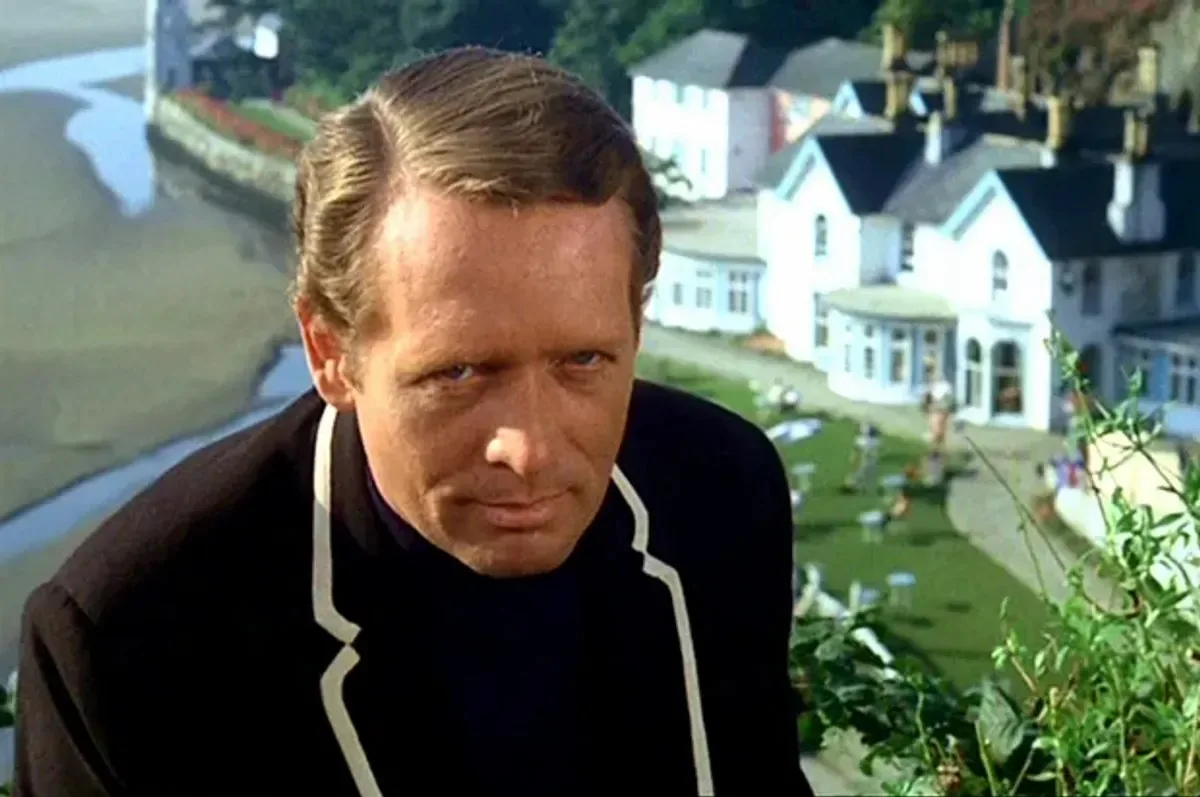 The image features a man with short, light brown hair styled neatly. He has a serious expression on his face, with a slight furrow in his brow and a focused gaze directed slightly off-camera. His clothing consists of a black jacket with a white trim along the collar and down the front, suggesting a formal or semi-formal style. The background reveals a scenic view of a coastal area, with a sandy beach visible on the left side of the image. 

In the background, there are several white buildings with dark roofs, likely residential or vacation homes, situated on a grassy area. The setting appears to be a sunny day, as indicated by the bright lighting and clear visibility. There are people in the background, some lounging on the grass, which adds a relaxed atmosphere to the scene. The overall composition suggests a blend of nature and architecture, with the man positioned in the foreground, creating a sense of depth.

No readable text, logos, or trademarks are present in the image. The focus remains on the individual and the picturesque environment surrounding him.