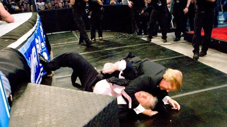The image depicts a wrestling scene occurring outside the ring, likely during a professional wrestling event. Two men are engaged in a physical altercation on the ground. 

1. **People and Actions**:
   - One man is on top of the other, appearing to grapple or wrestle. The man on the bottom is wearing a light pink dress shirt and black pants, while the man on top is dressed in a black suit with a white shirt.
   - The man in the pink shirt has a facial expression that suggests he is either surprised or in distress, while the man in the black suit appears to be exerting force.
   - Both individuals are on a black mat that covers the floor, indicating they are outside the wrestling ring.

2. **Clothing**:
   - The man in the pink shirt has rolled up sleeves and is wearing a black tie. His hair is dark and styled.
   - The man in the black suit has slicked-back hair and is wearing a black tie as well. His suit appears well-fitted.

3. **Setting**:
   - The background shows a wrestling ring with a blue and white banner, likely featuring a logo or branding associated with the wrestling promotion.
   - There are several individuals in black clothing visible in the background, possibly security personnel or event staff, observing the altercation.
   - The audience is partially visible, with many people in the background watching the event, indicating a lively atmosphere.

4. **Environmental Context**:
   - The scene is set in a large indoor arena, typical for wrestling events, with bright lights illuminating the ring and surrounding area.
   - The flooring is a dark, padded surface, which is common in wrestling venues to provide safety during matches.

Overall, the image captures a dynamic moment in a wrestling event, characterized by physical engagement between two individuals in formal attire, set against the backdrop of an audience and a wrestling ring.